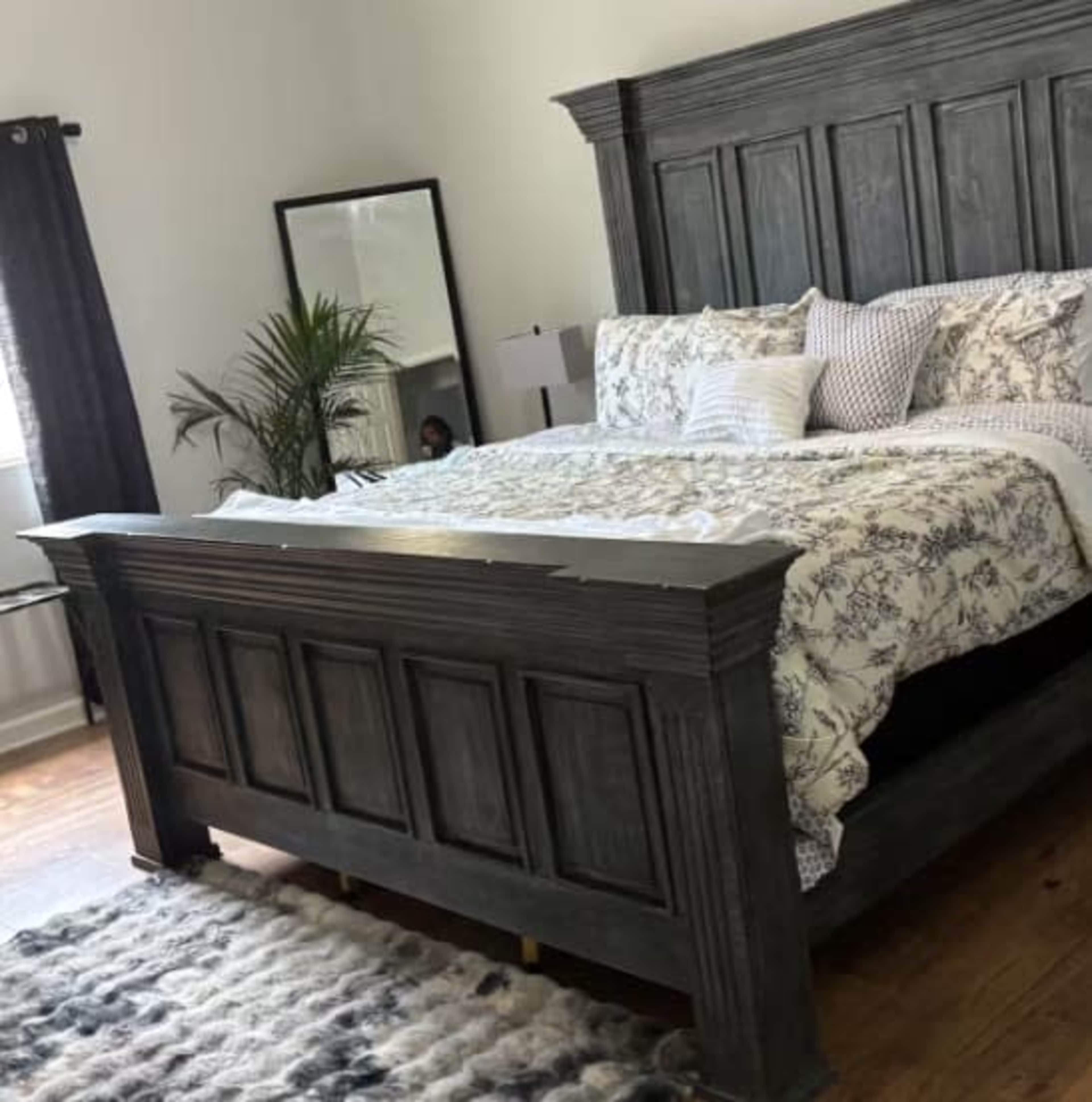 The image shows a spacious bedroom featuring a large gray wooden bed with a patterned comforter, flanked by a mirrored dresser and a plant in the corner.