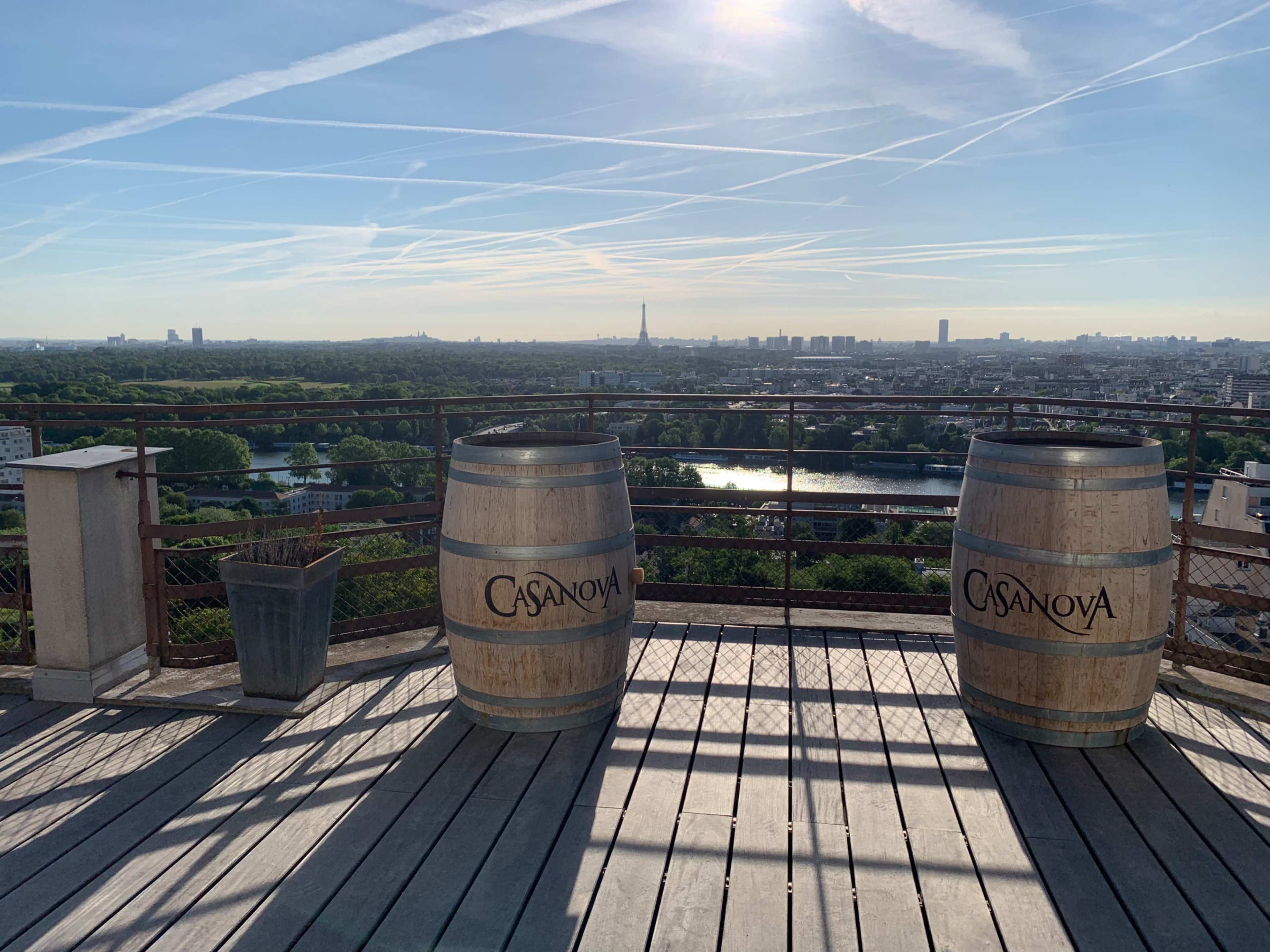 A rooftop terrace with two wooden barrels labeled "Casanova" overlooking a cityscape featuring the Eiffel Tower and a river.