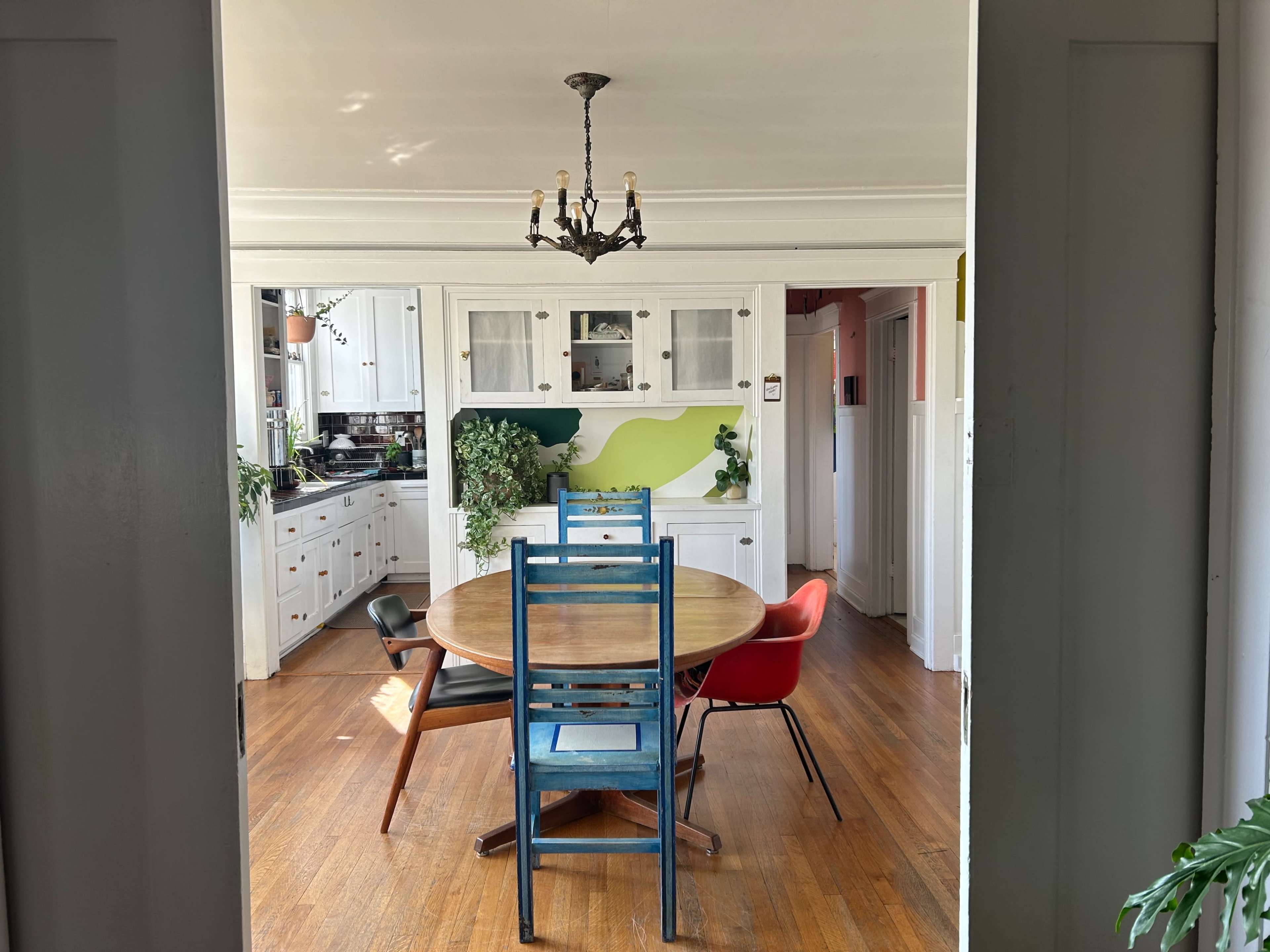 A dining room features a round wooden table surrounded by four chairs, with a view of a kitchen in the background.