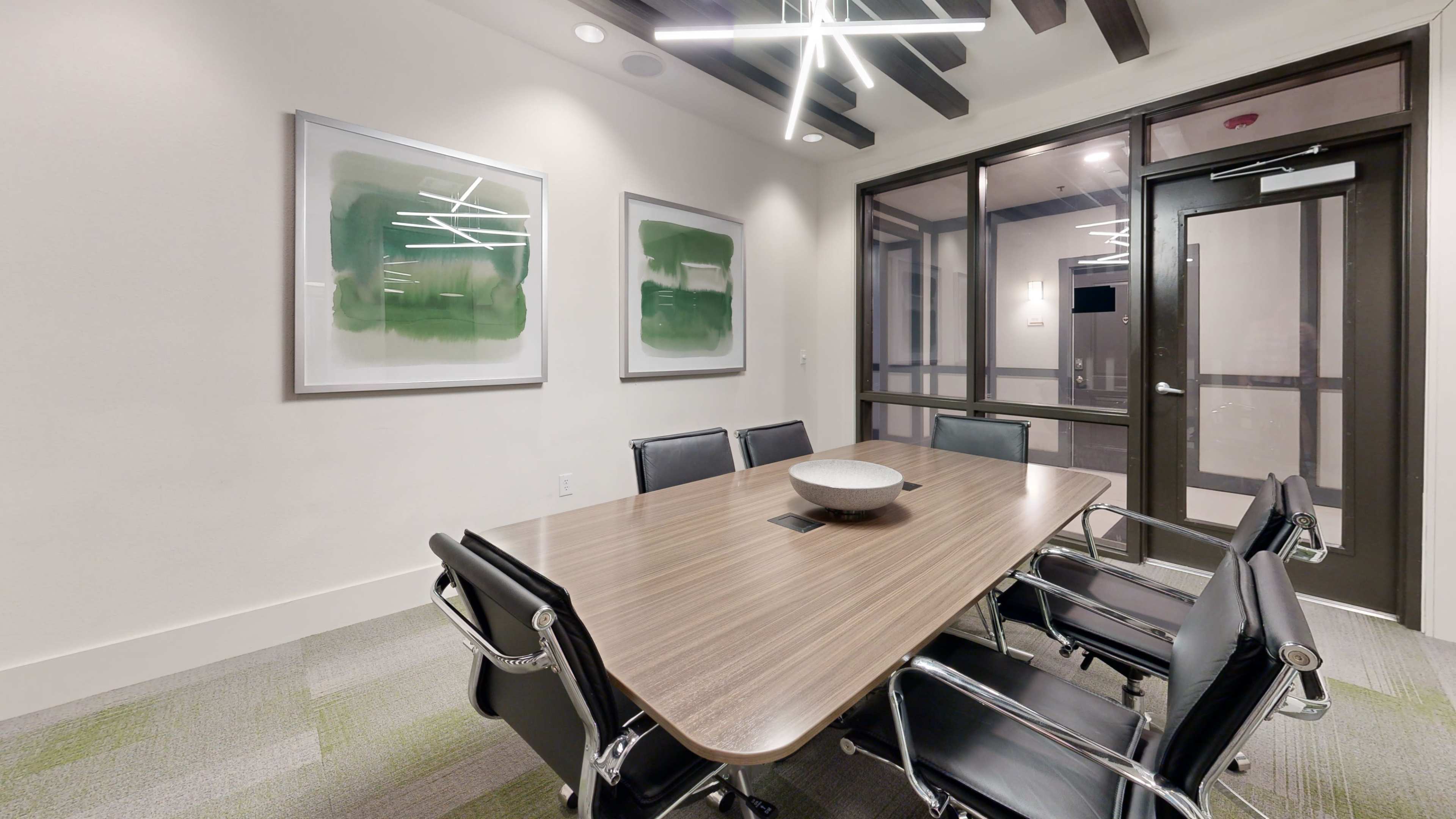 The image shows a modern conference room with a long wooden table surrounded by black chairs, framed abstract artwork on the walls, and glass doors.