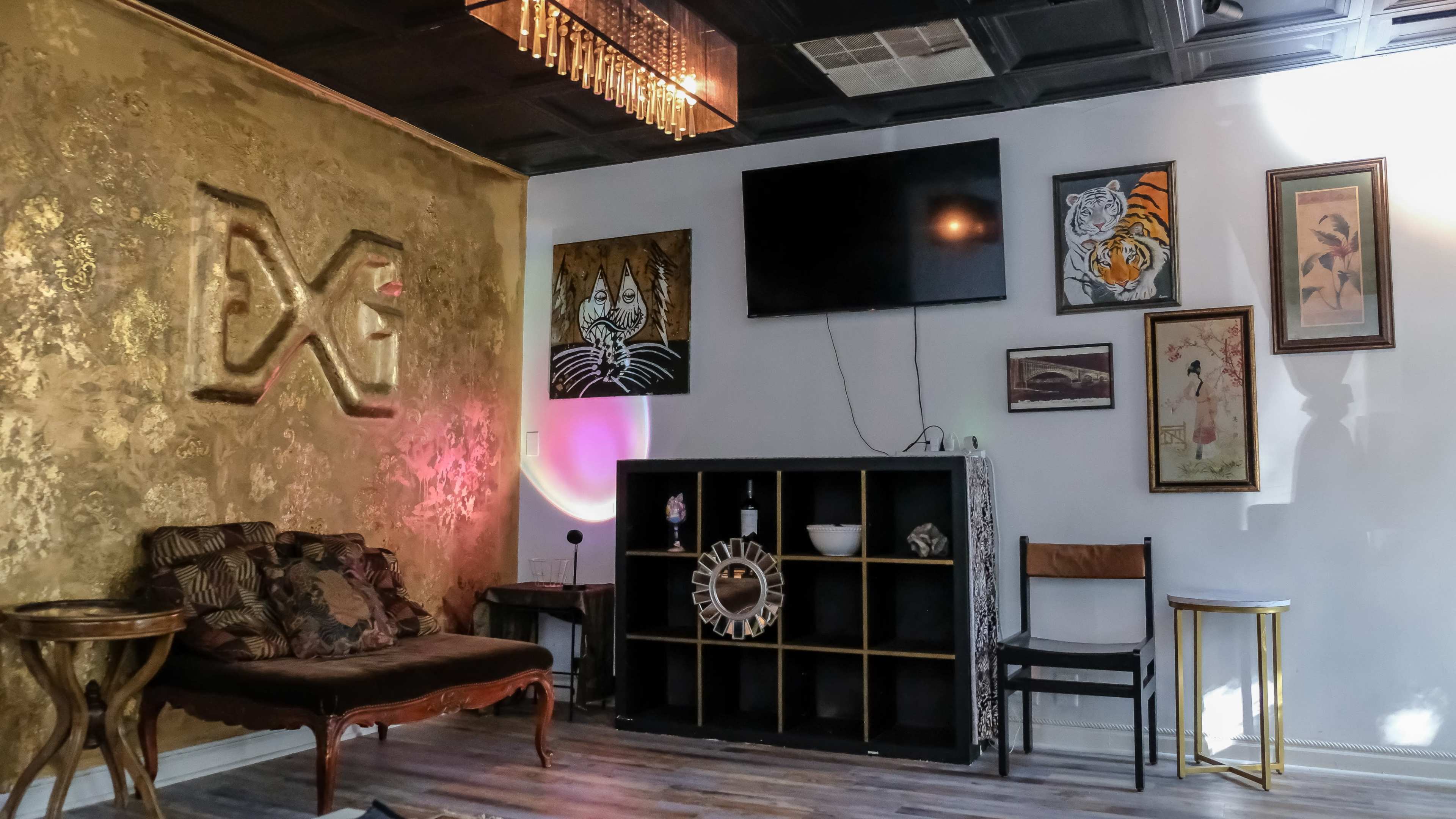 The image shows a modern room with golden walls, a television mounted on the wall, various framed artworks, and a decorative shelf holding items alongside a couch and chairs.