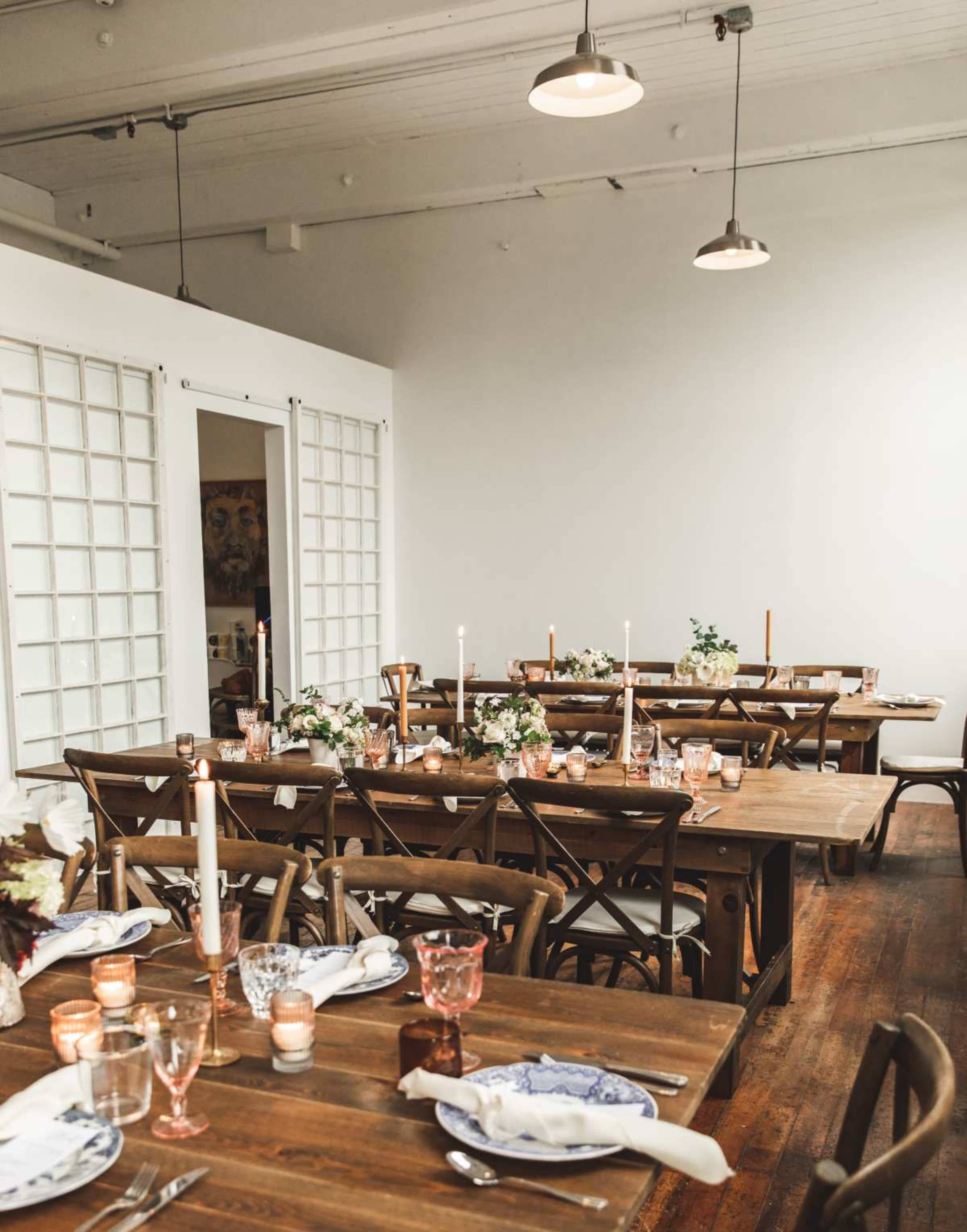 The image shows a dining area with several wooden tables set for a meal, featuring candle holders, floral centerpieces, and tableware.