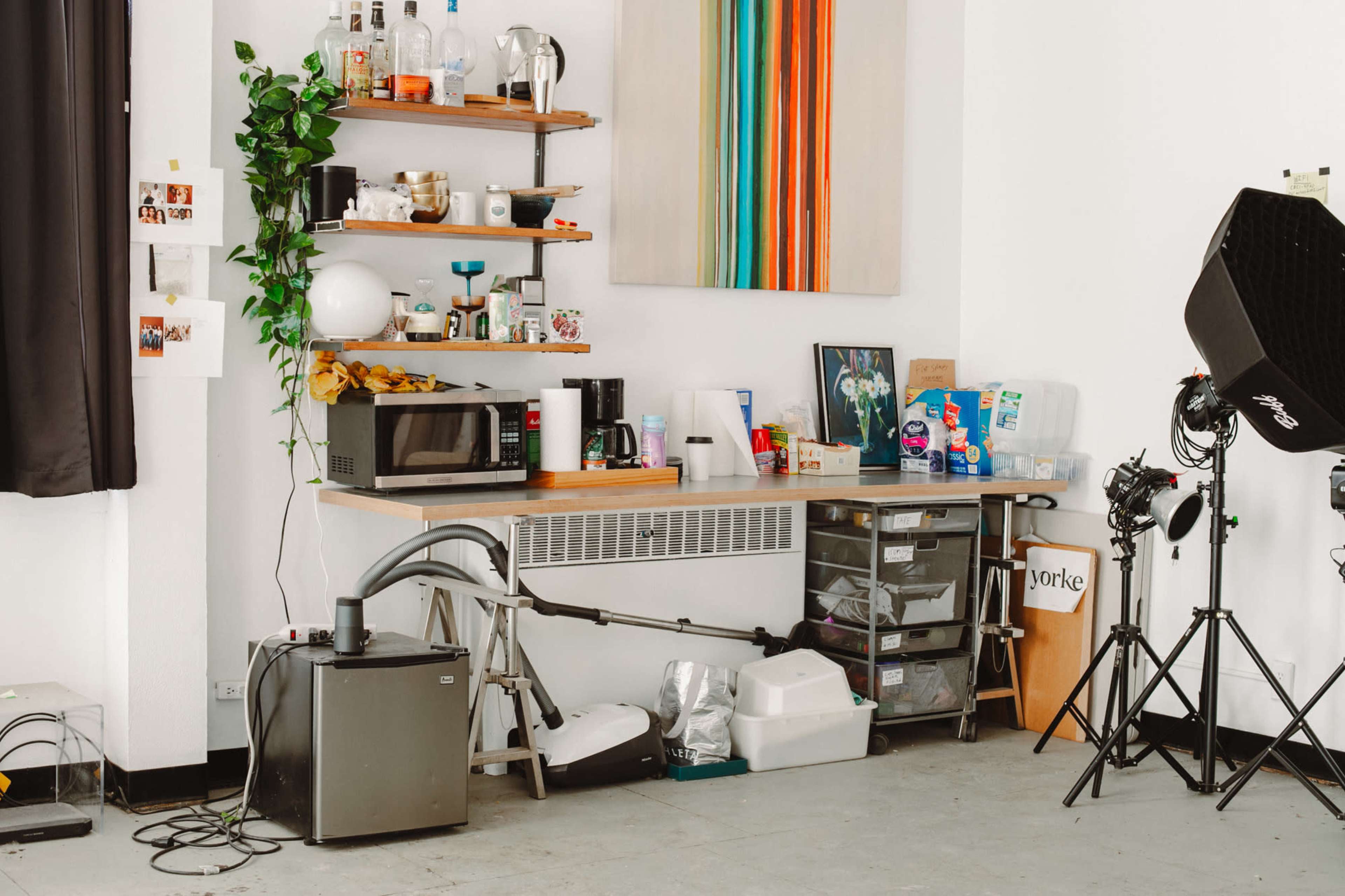 The image shows a workspace with a large wooden table, a microwave, shelving with various bottles and plants, and several storage bins along with photography equipment.