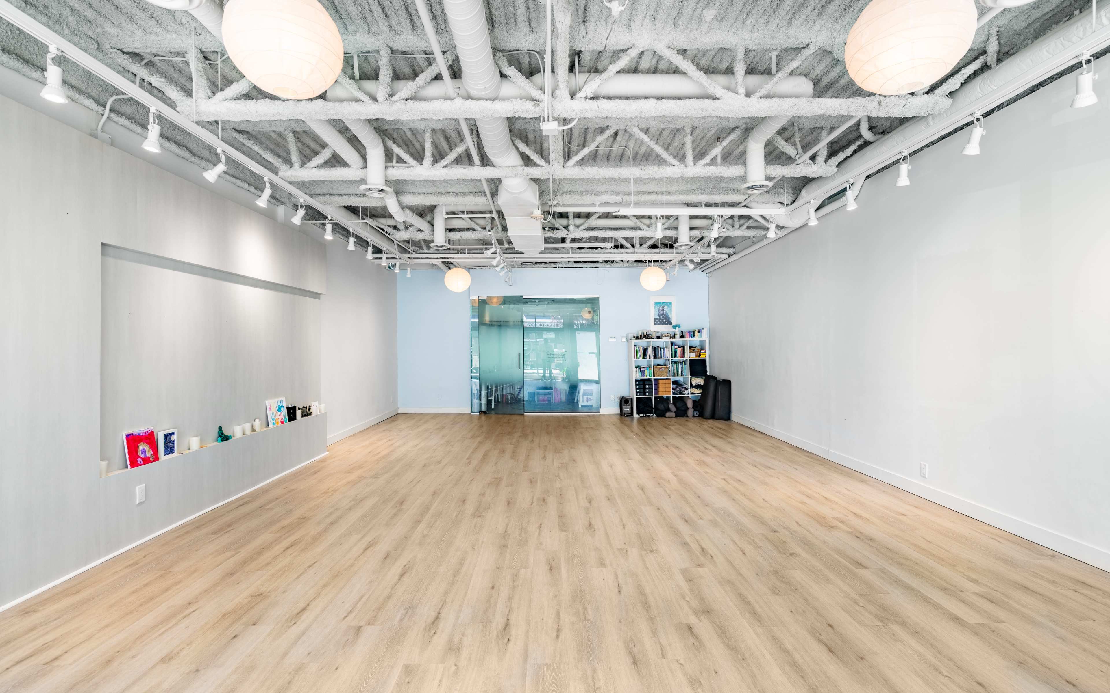 A spacious, well-lit interior with wooden flooring, white walls, and a glass entrance, featuring shelves and decorative items along one side.