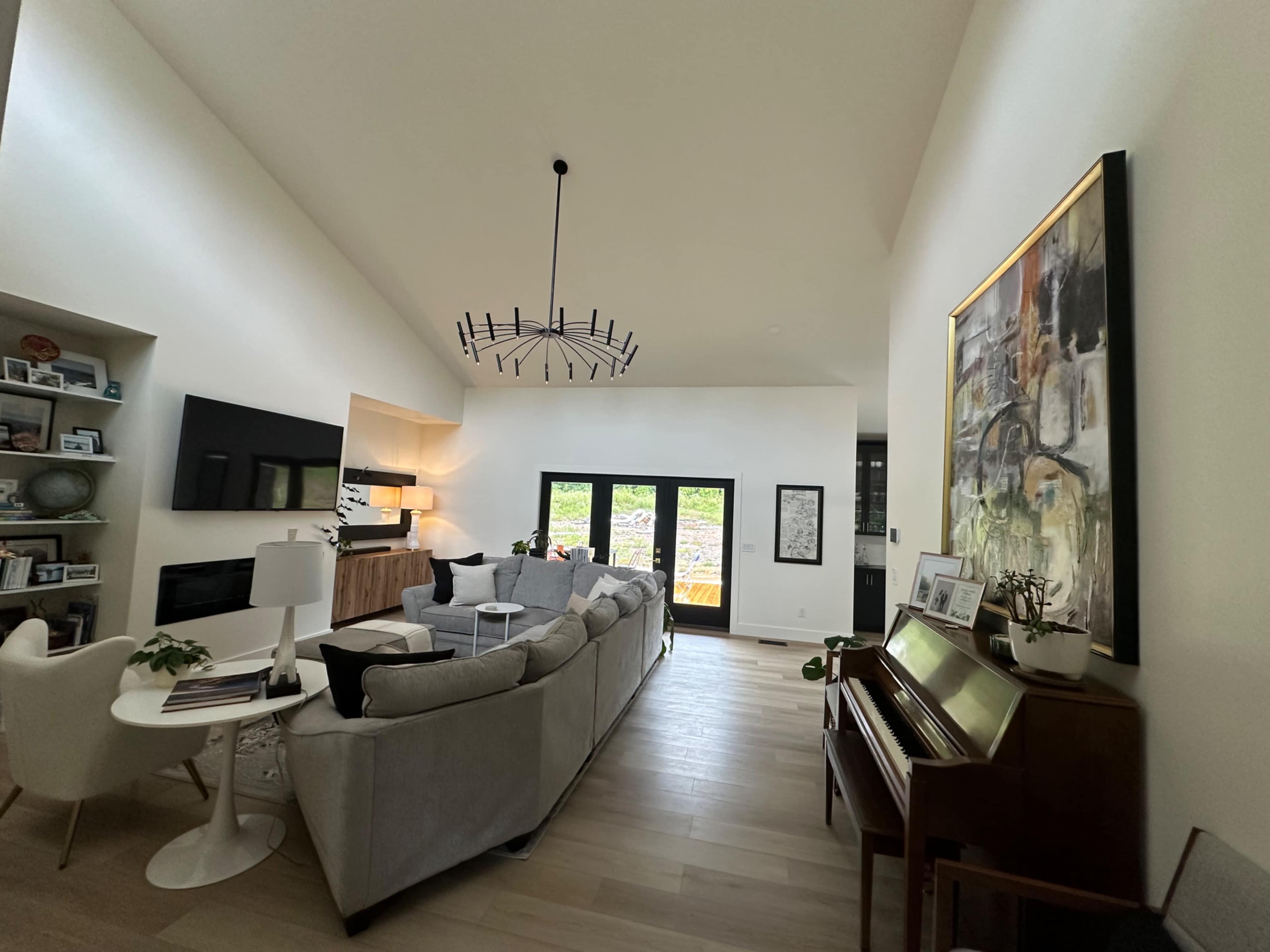 A spacious living room features a gray sectional sofa, a wooden piano, and large windows that open to a view of greenery.