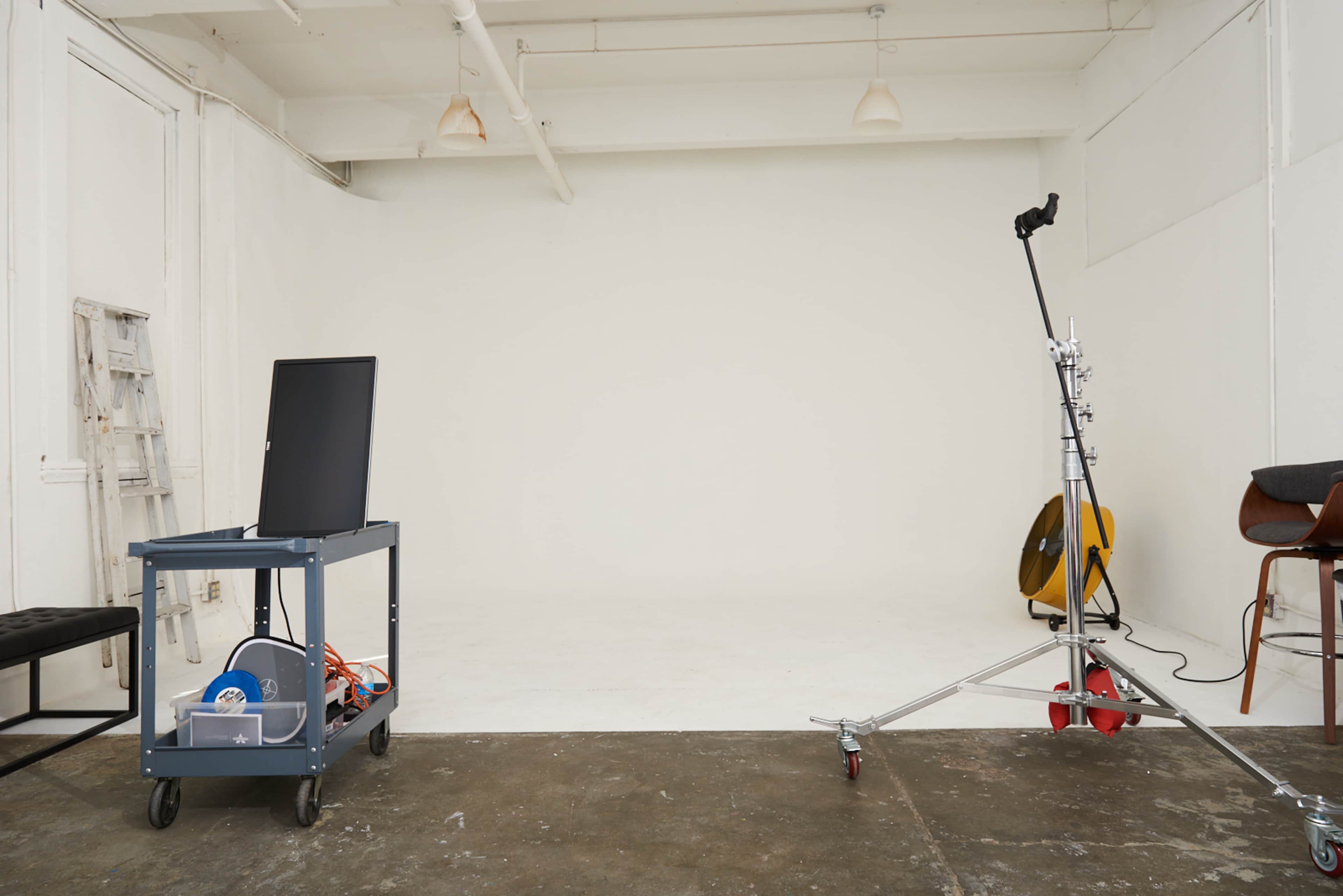 A modern studio space features a blank white wall, a cart with equipment, a light stand, and a chair in the corner.