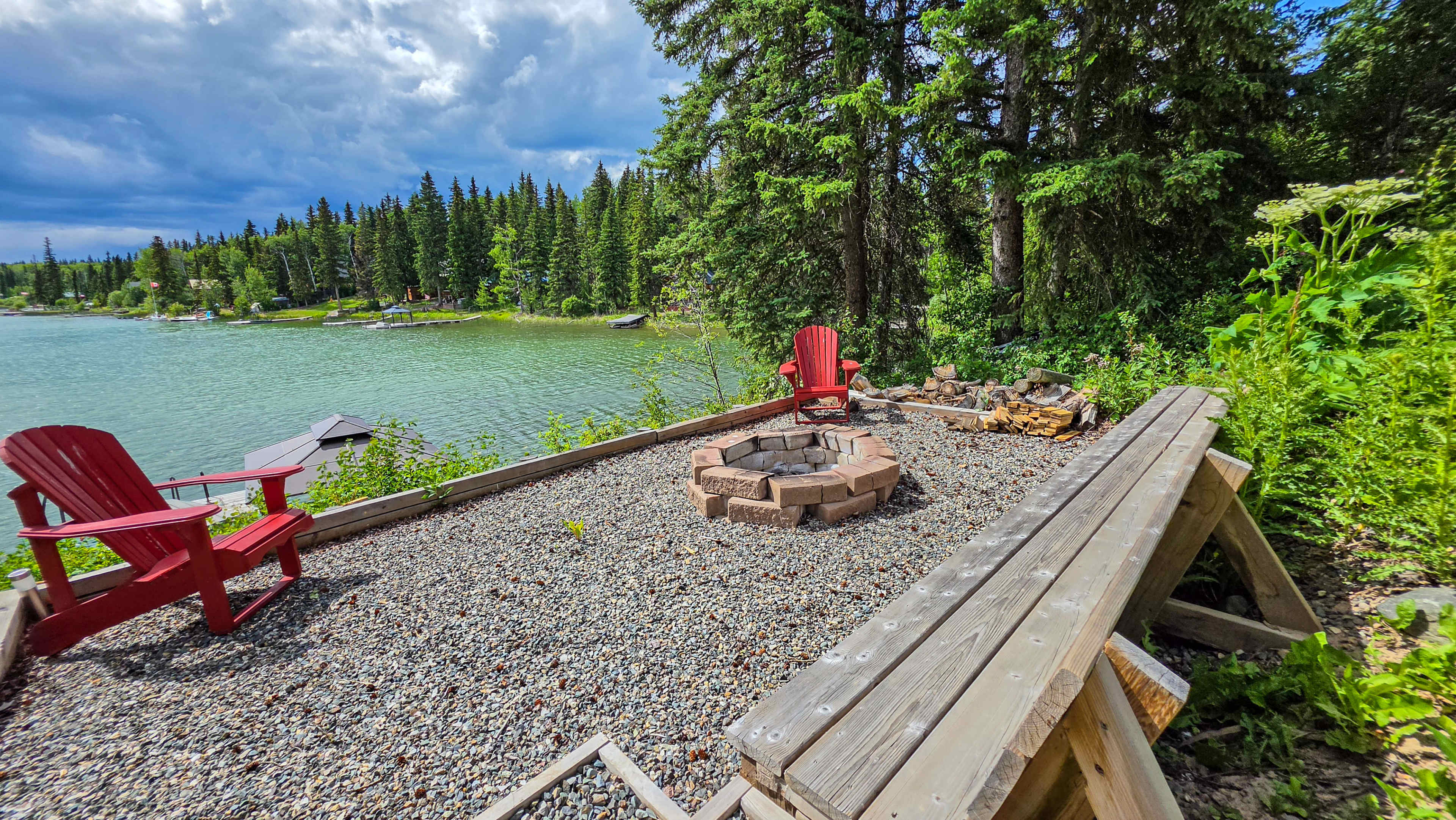 The image shows a lakeside seating area with two red chairs surrounding a stone fire pit, overlooking a water body surrounded by trees.