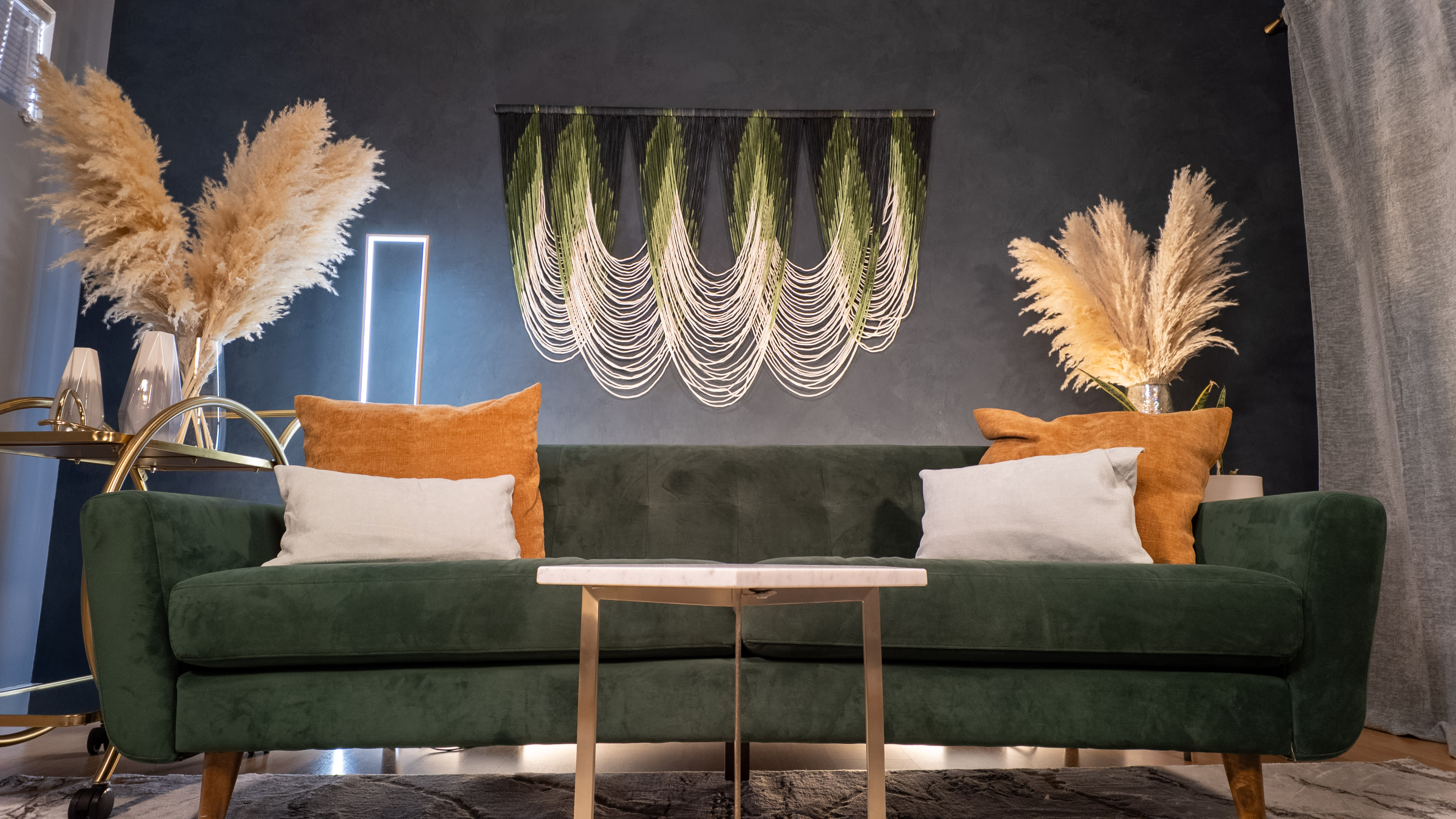 A green sofa is positioned in front of a dark wall, adorned with a decorative piece made of strings and greenery, with two pillows and a marble-topped table placed in front.