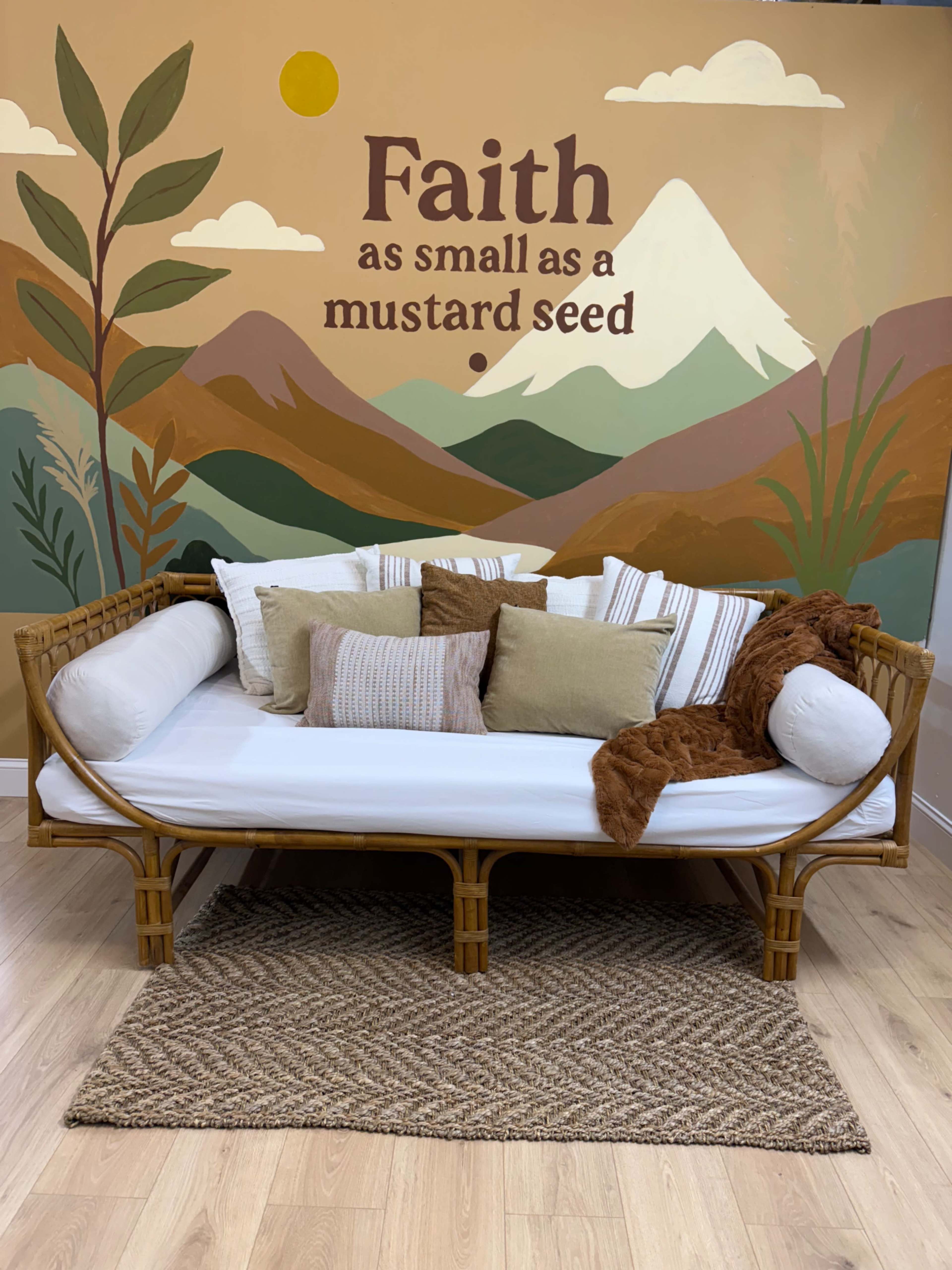 A daybed with decorative pillows sits against a mural featuring mountains and foliage, accompanied by the text "Faith as small as a mustard seed."