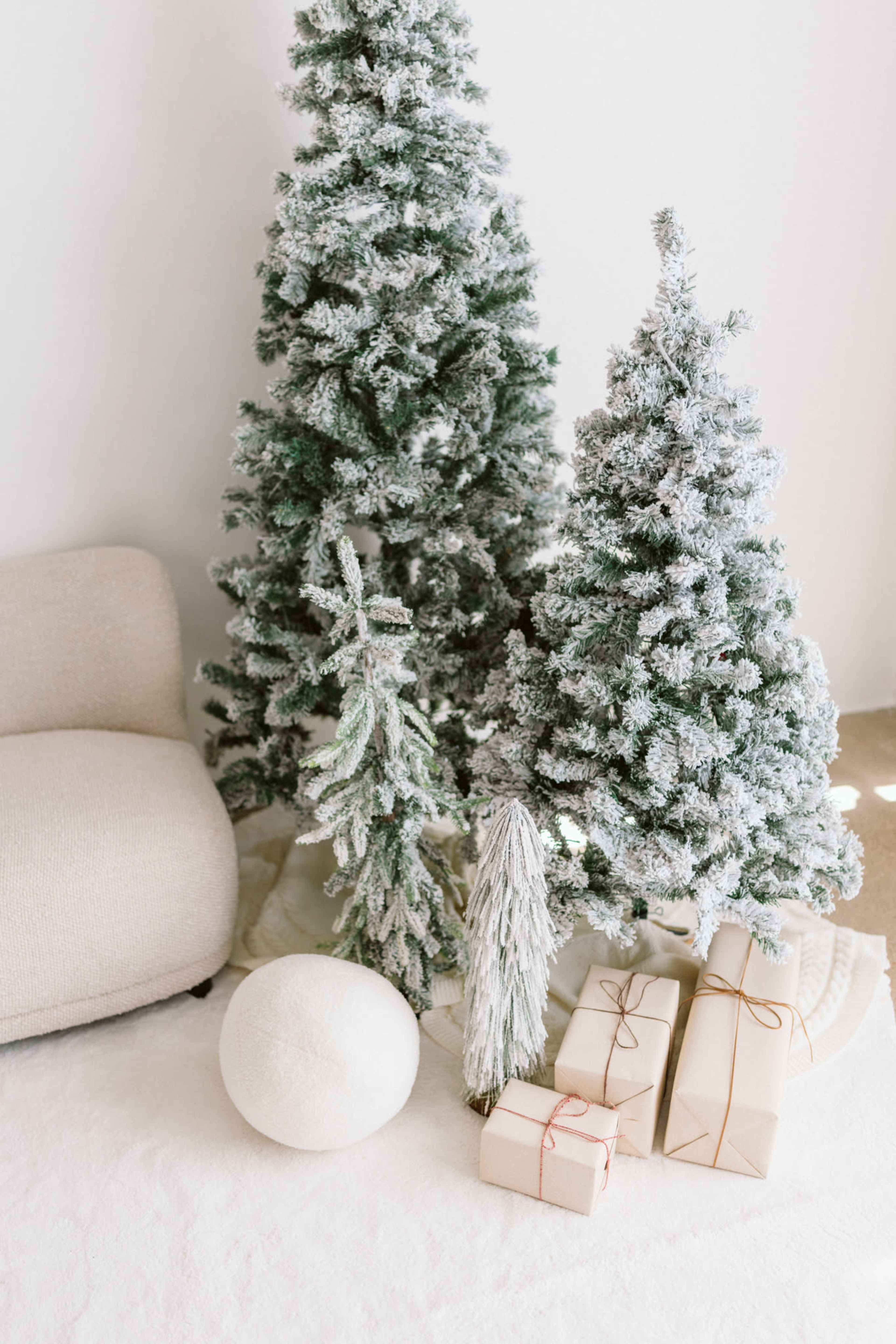 The image features two frosted Christmas trees beside a cozy beige chair, with neatly wrapped gifts placed on a soft rug.