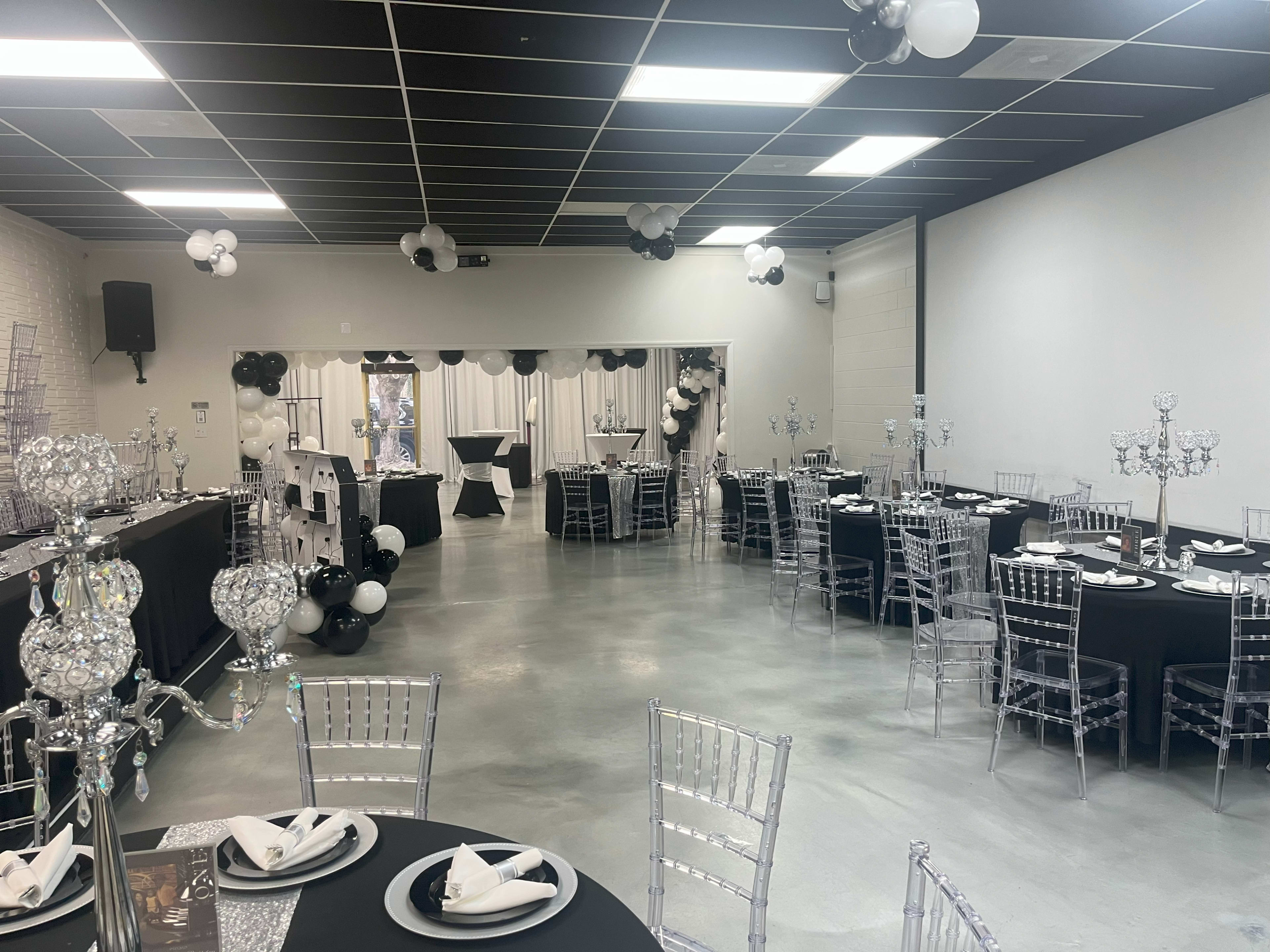 The image shows a spacious event hall decorated with black and white theme elements, featuring tables with black tablecloths, crystal candelabras, and balloons.