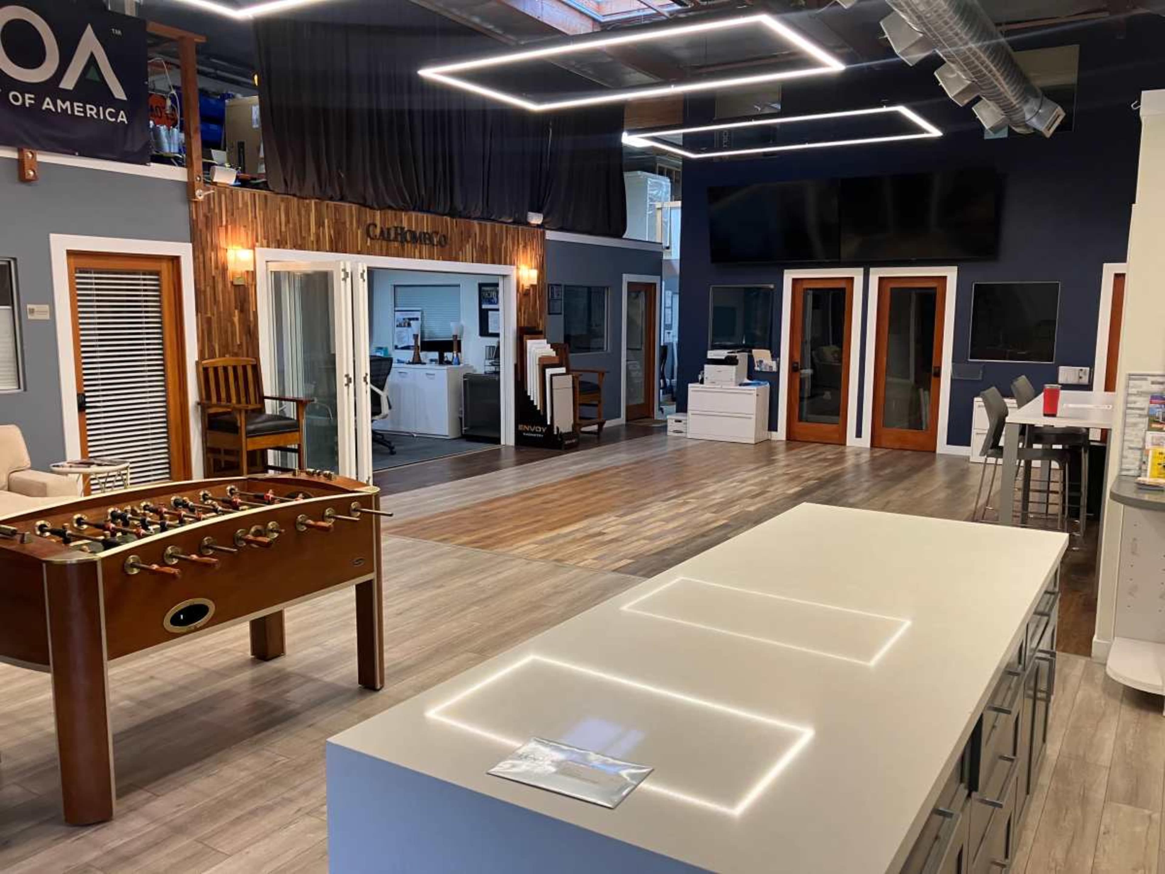 The image shows a modern showroom featuring a foosball table in the foreground and display areas with furniture and office setups in the background.