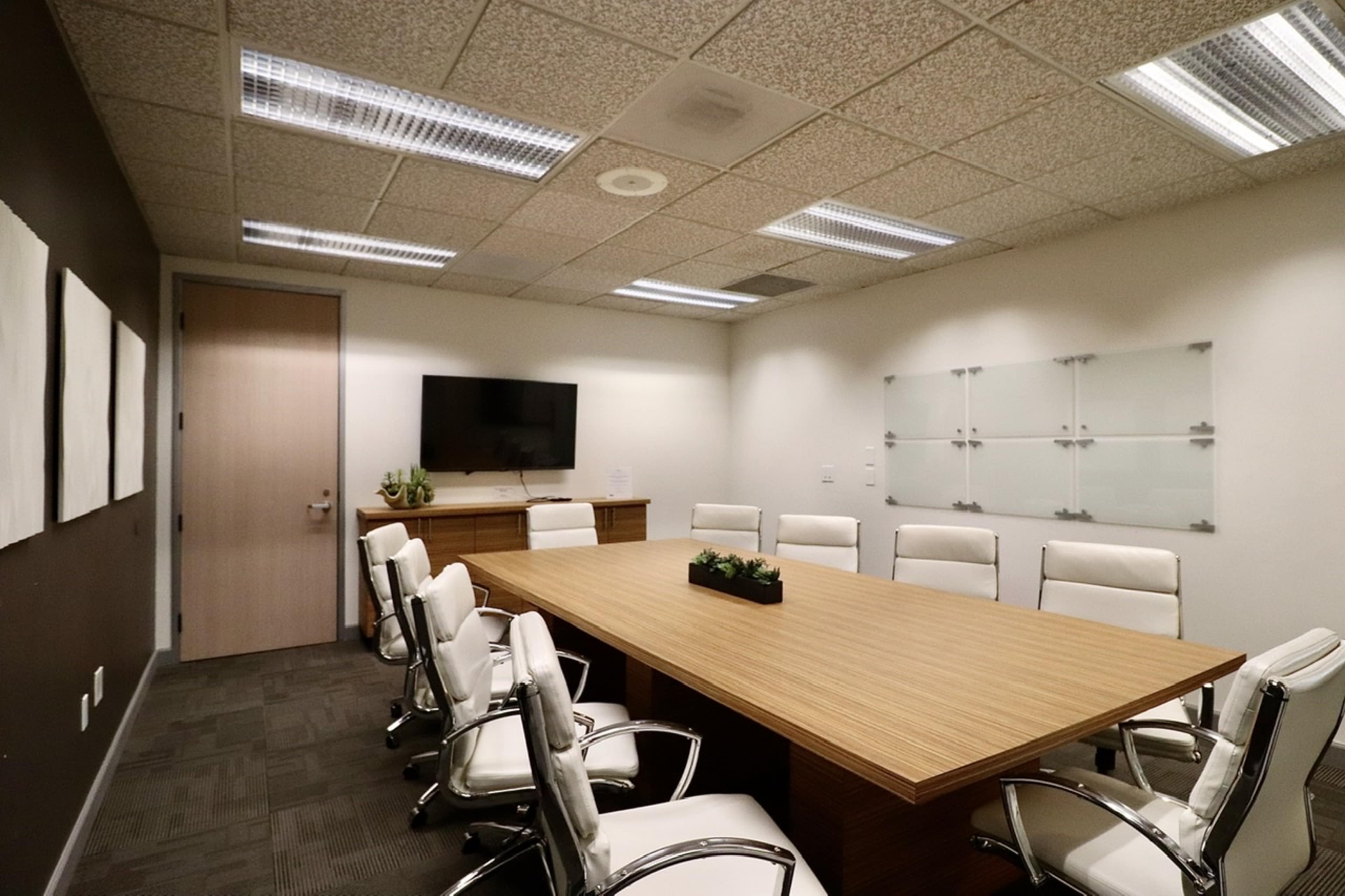 A modern conference room features a large wooden table surrounded by white chairs, with a television and glass boards on the walls.