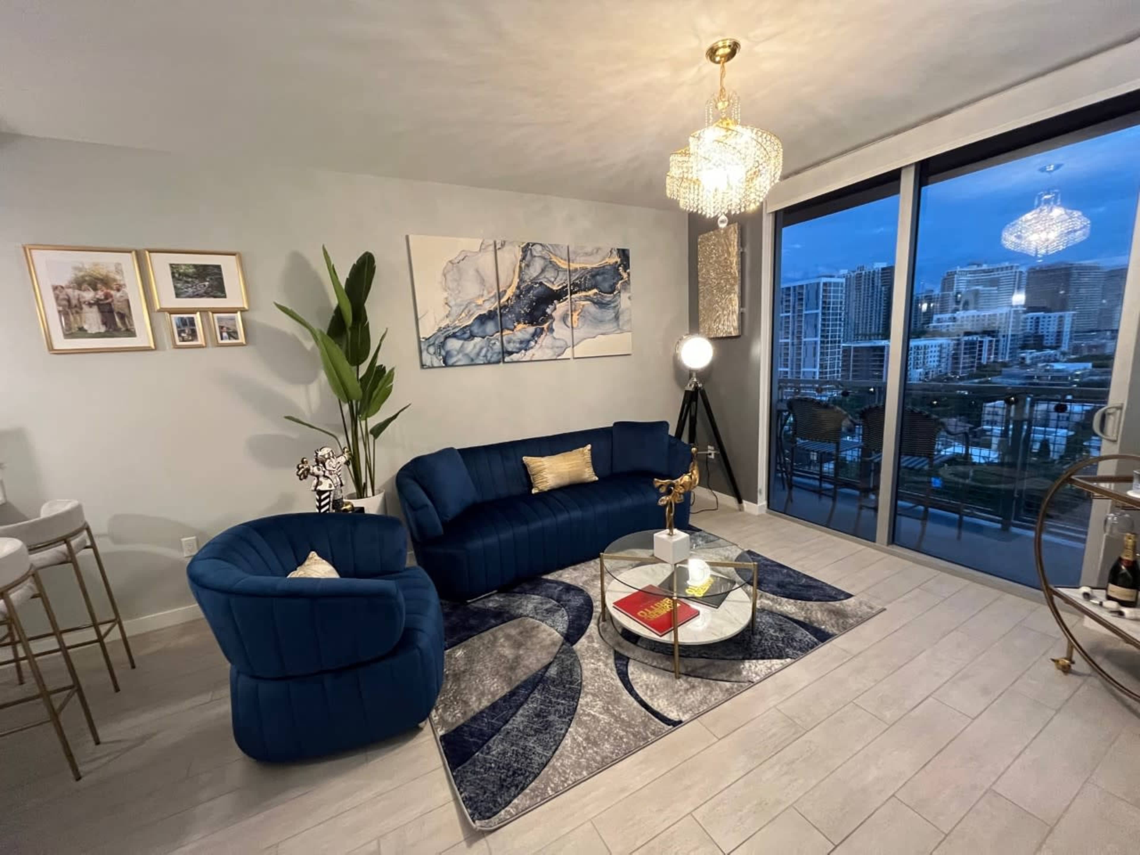 A stylish living room features a blue velvet sofa, a round glass coffee table, and large windows overlooking a cityscape.