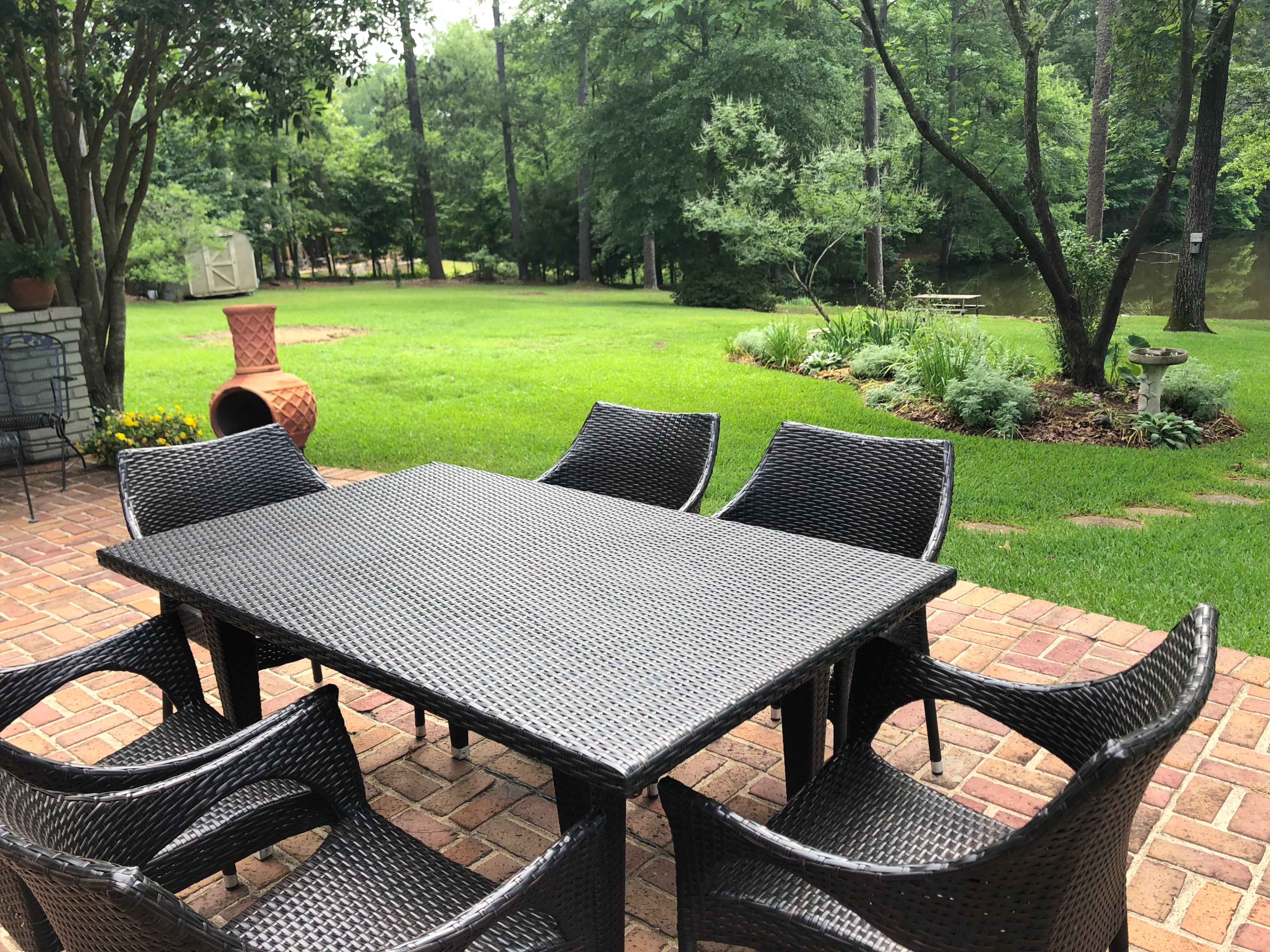 A wicker dining table with six chairs is positioned on a brick patio overlooking a grassy yard and a pond surrounded by trees.