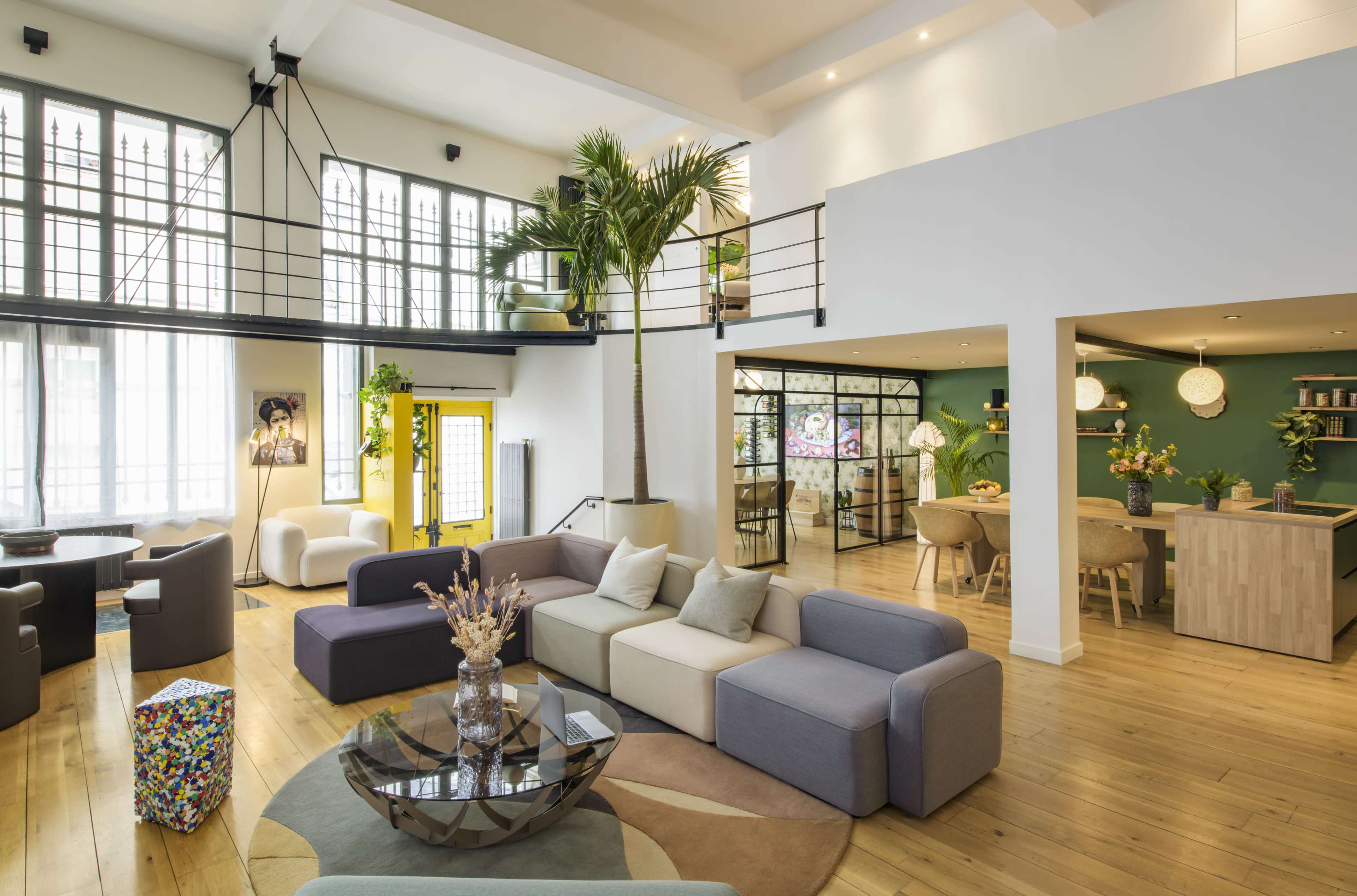 The image shows a modern, open-plan living area with a mix of seating options and a vibrant color scheme, featuring large windows and a decorative indoor plant.