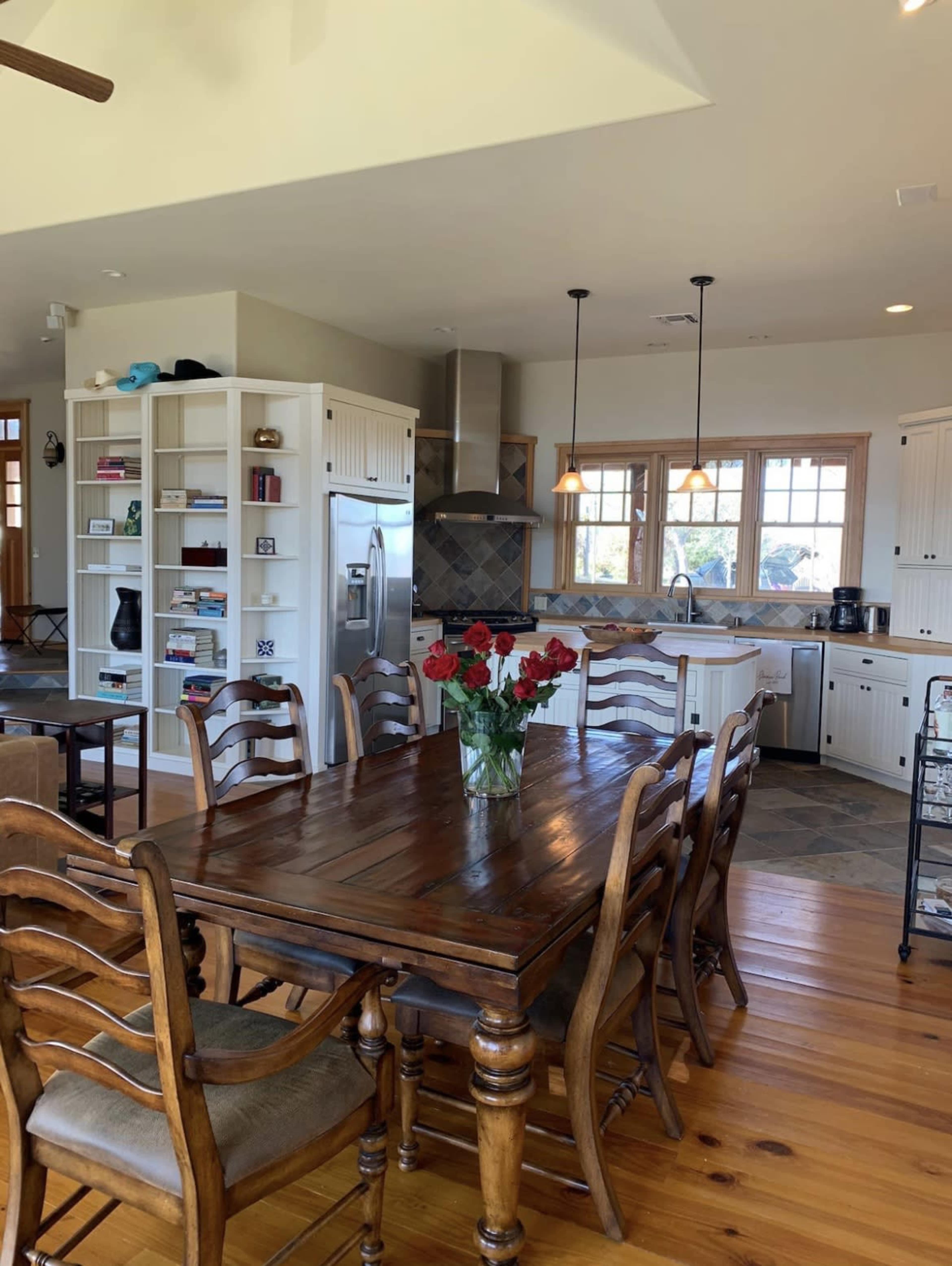 A spacious kitchen and dining area featuring a wooden dining table with chairs, a vase of red roses, white cabinets, and a stainless steel refrigerator.
