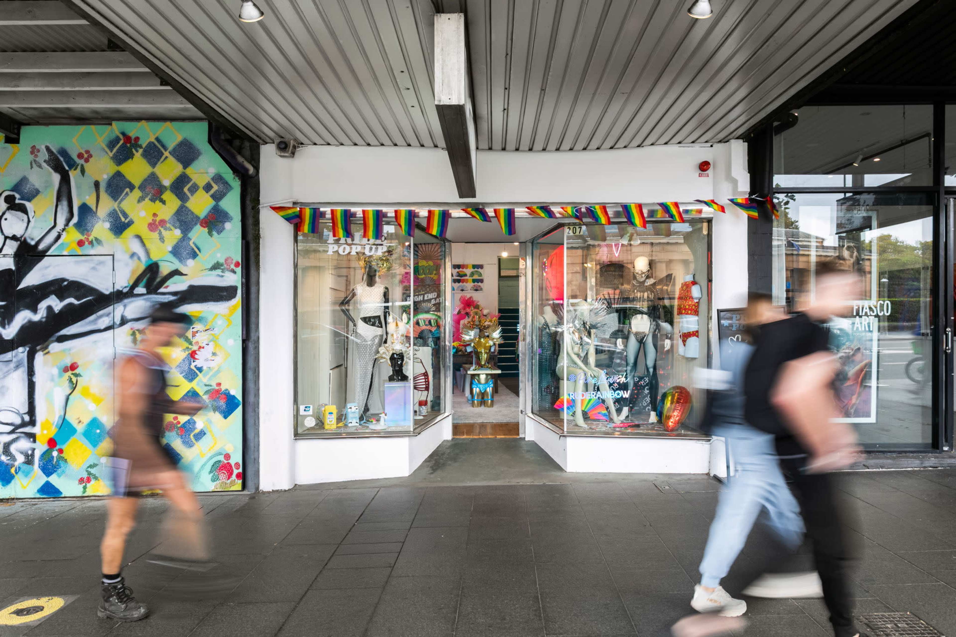 The image shows a storefront adorned with colorful decorations and a window display featuring various items, while pedestrians walk by on the sidewalk.