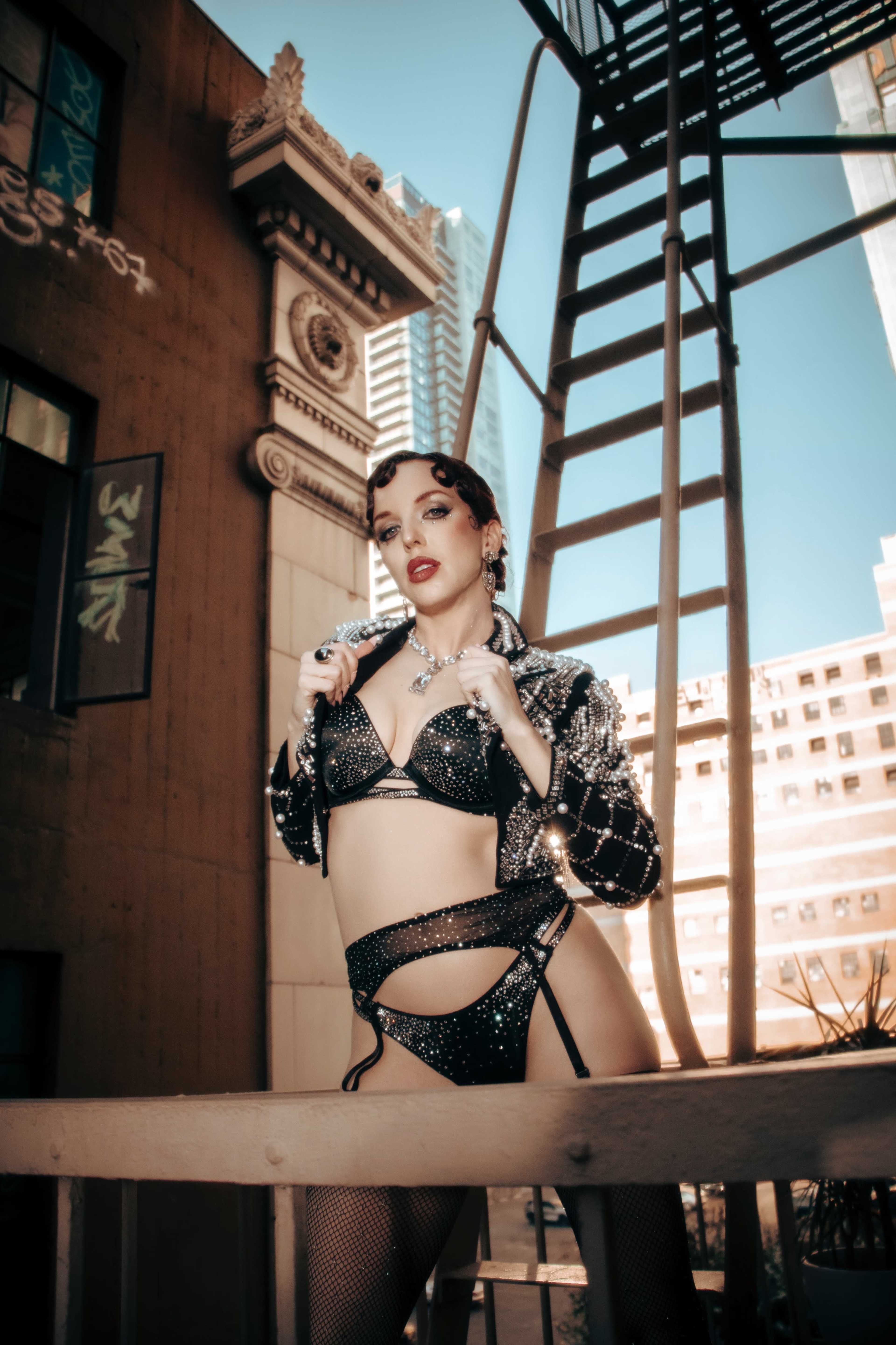 A person in a sparkling black outfit poses on a balcony with a fire escape and urban buildings in the background.