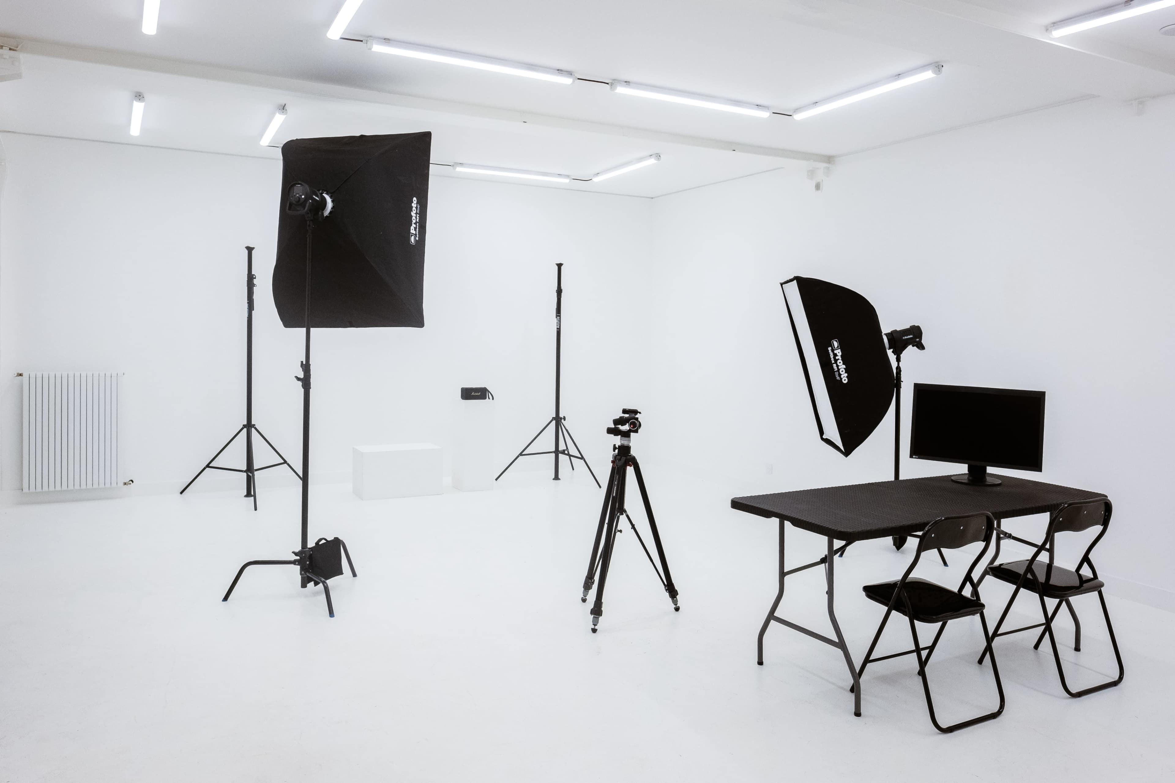 A photography studio with lighting equipment, a camera on a tripod, a table with chairs, and a monitor, all arranged in a minimalist white space.