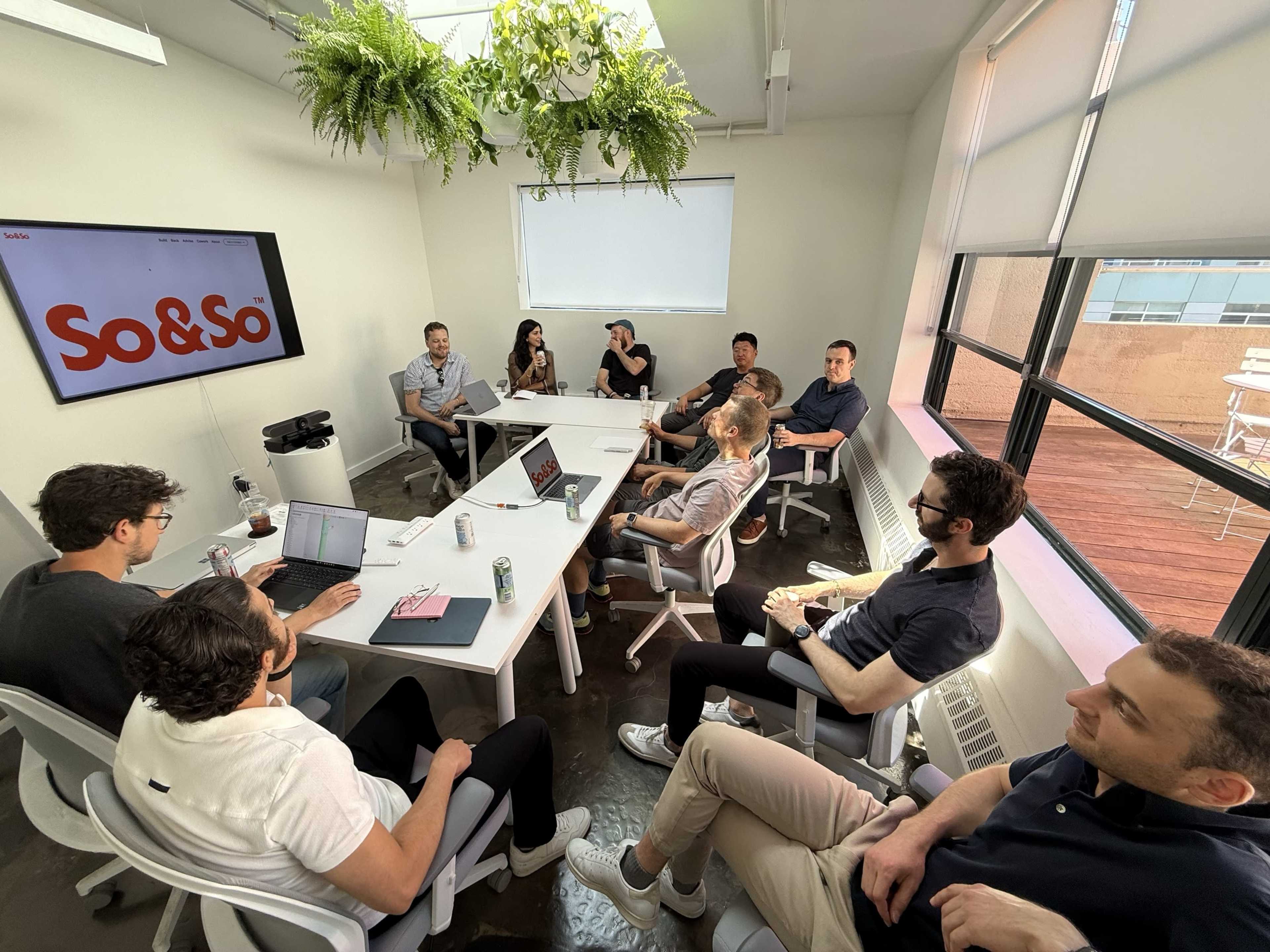 A group of ten people is seated around a large white conference table in a brightly lit meeting room, with a presentation screen displaying "So&So" in the background.