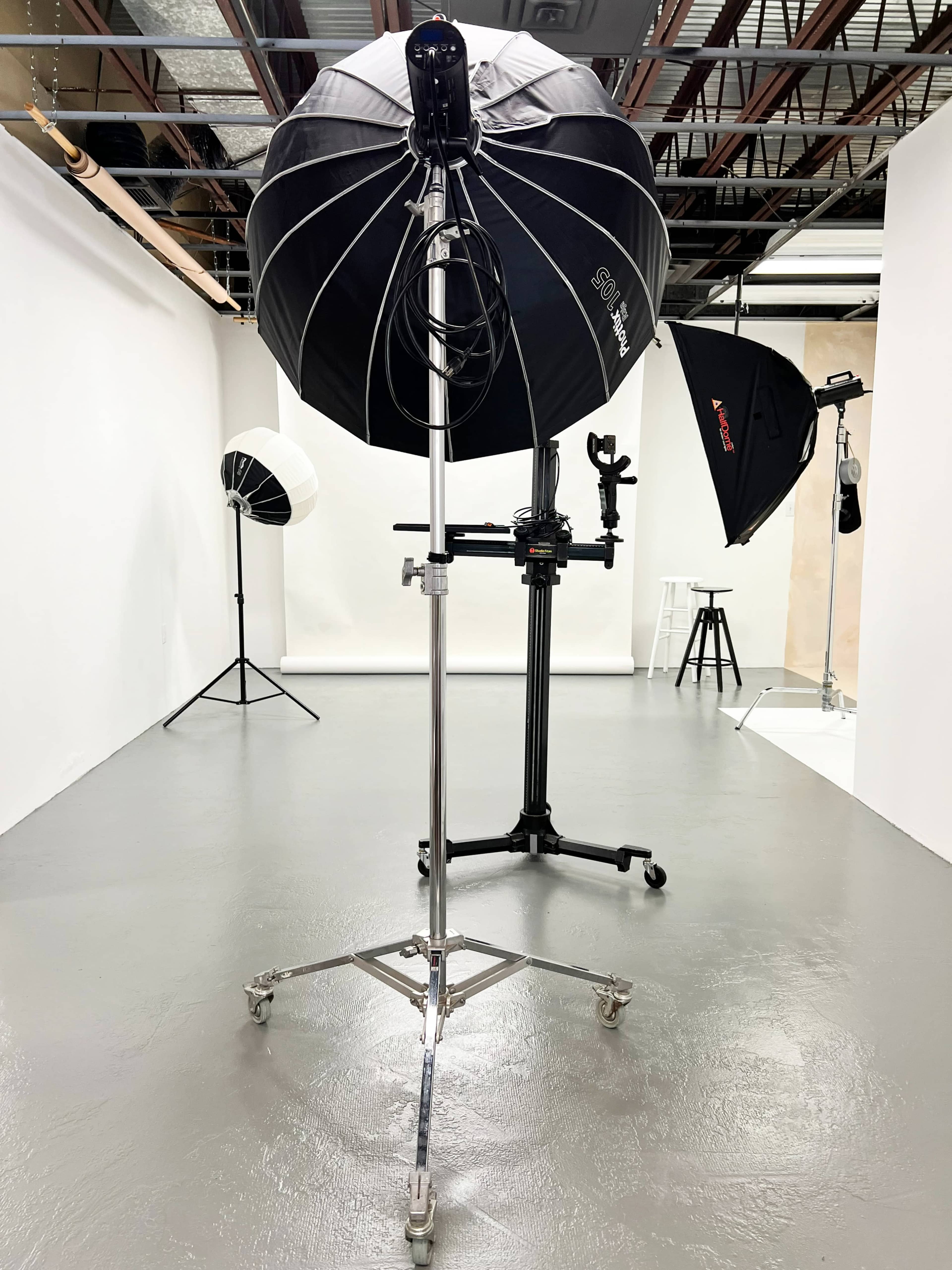 A studio lighting setup includes a large black umbrella softbox on a stand, with additional lighting equipment and a stool visible in a spacious, well-lit room.