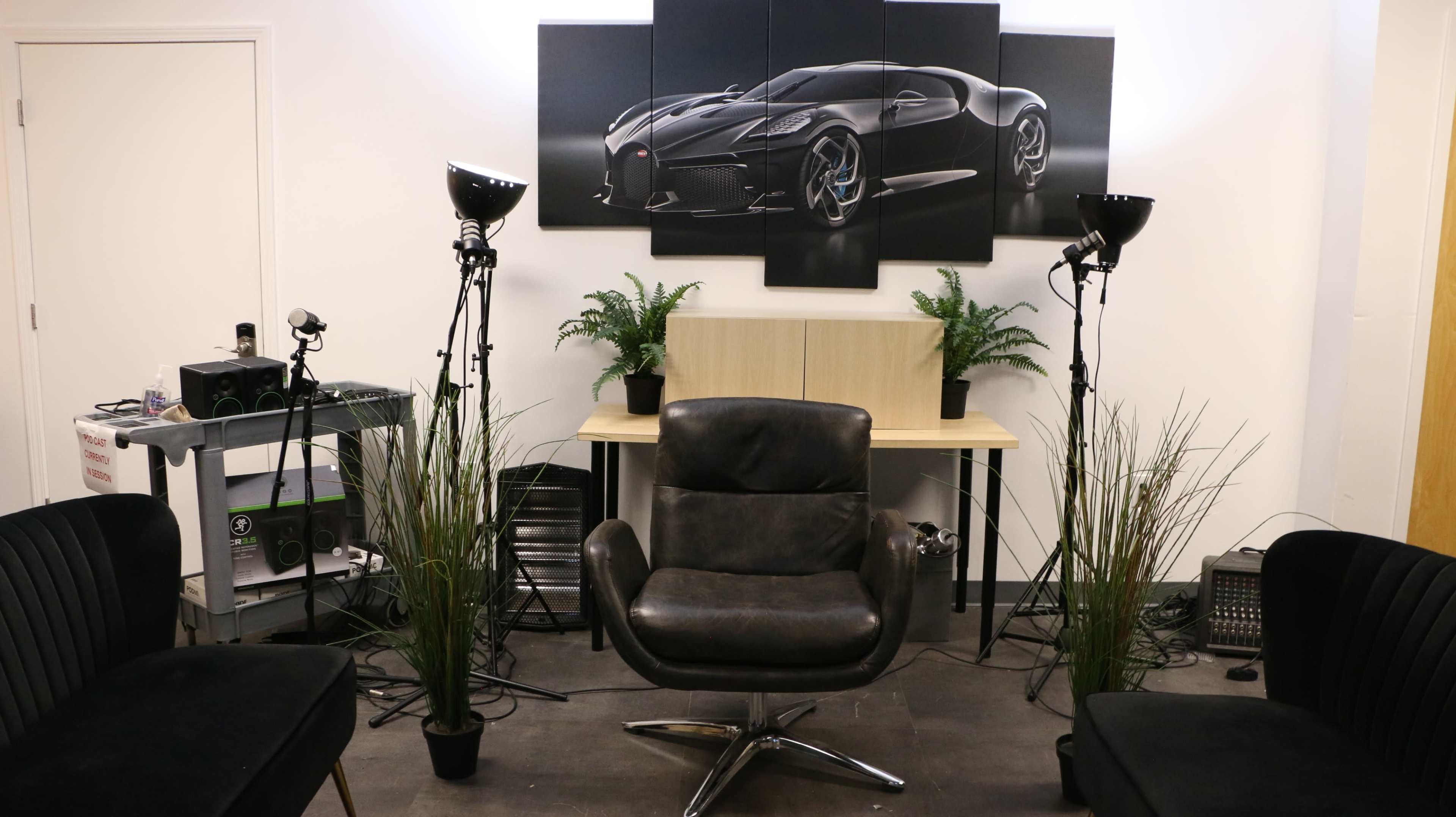 The image depicts a modern lounge area with a central black armchair, surrounded by potted plants, and illuminated by floor lamps, with a large car-themed artwork mounted on the wall behind.