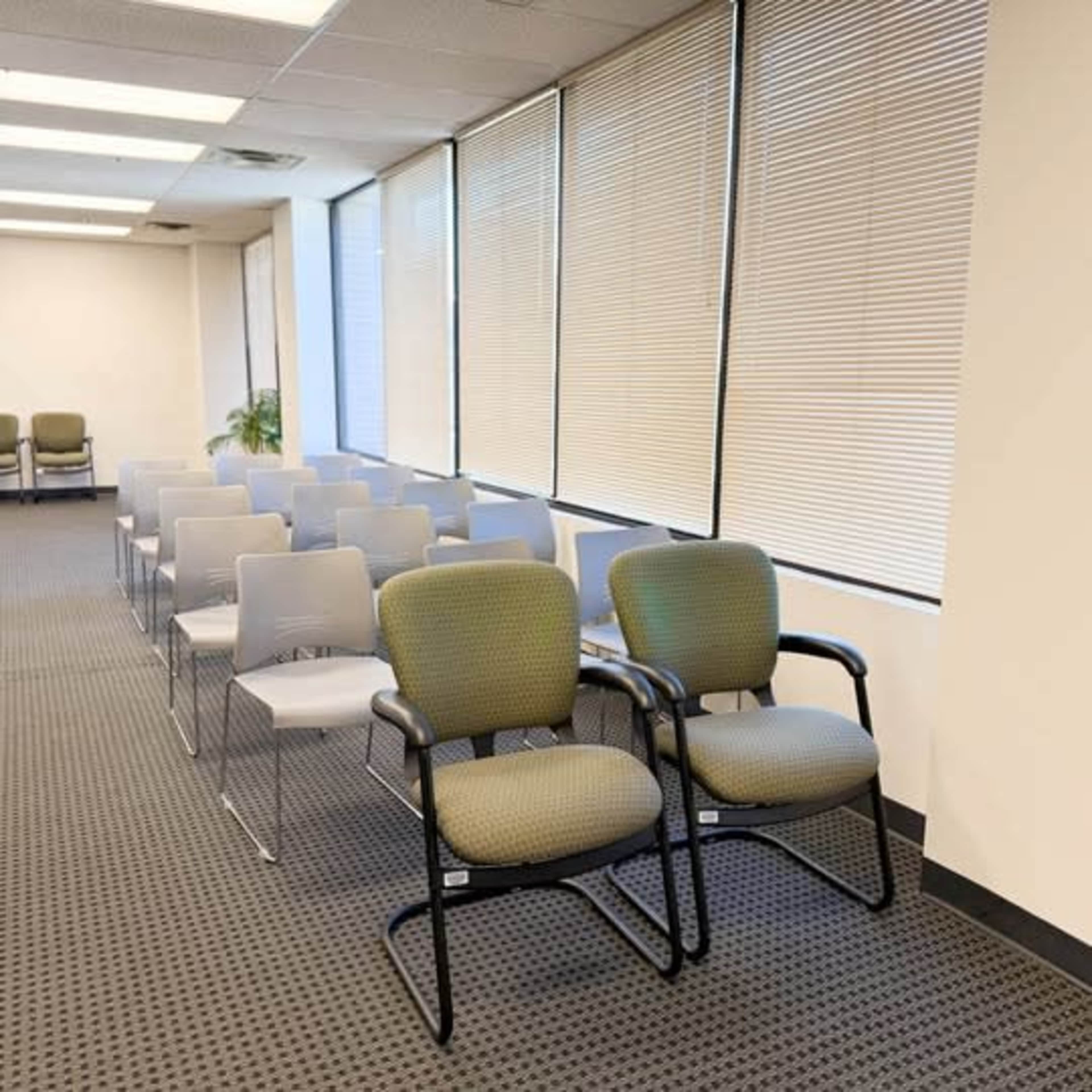 The image shows a row of empty chairs in a waiting area with large windows covered by blinds.