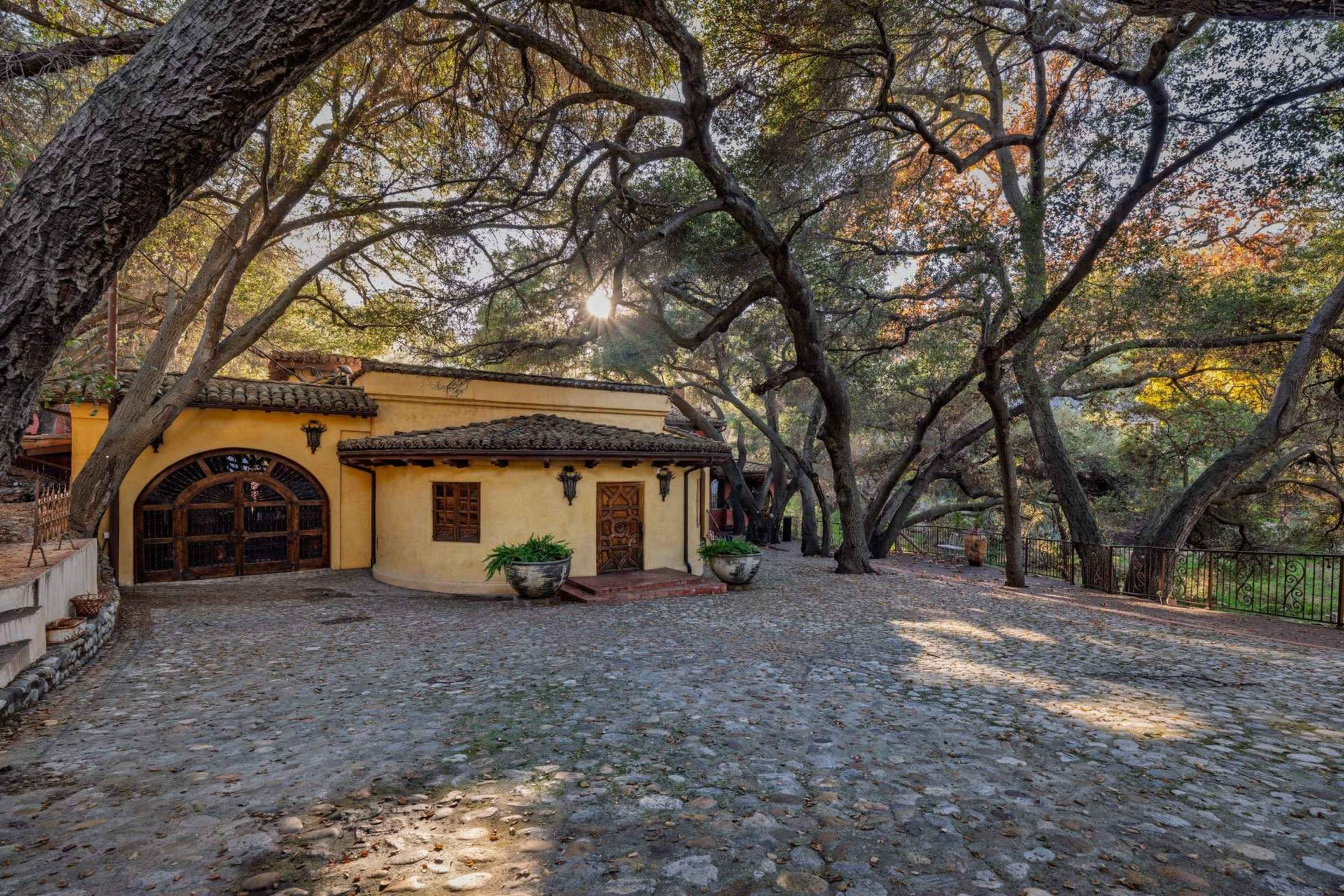 A stone pathway leads to a round, yellow building surrounded by large trees and a peaceful outdoor area.
