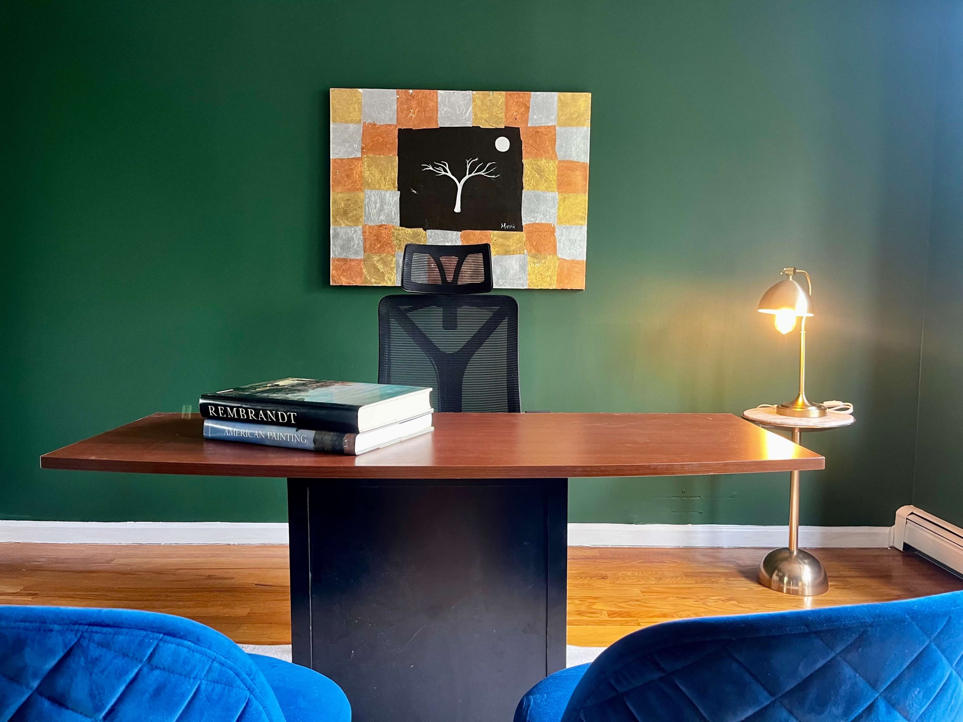 A minimalist office features a dark green wall, a wooden desk with two stacked art books, an ergonomic chair, and a lamp beside a colorful abstract painting.