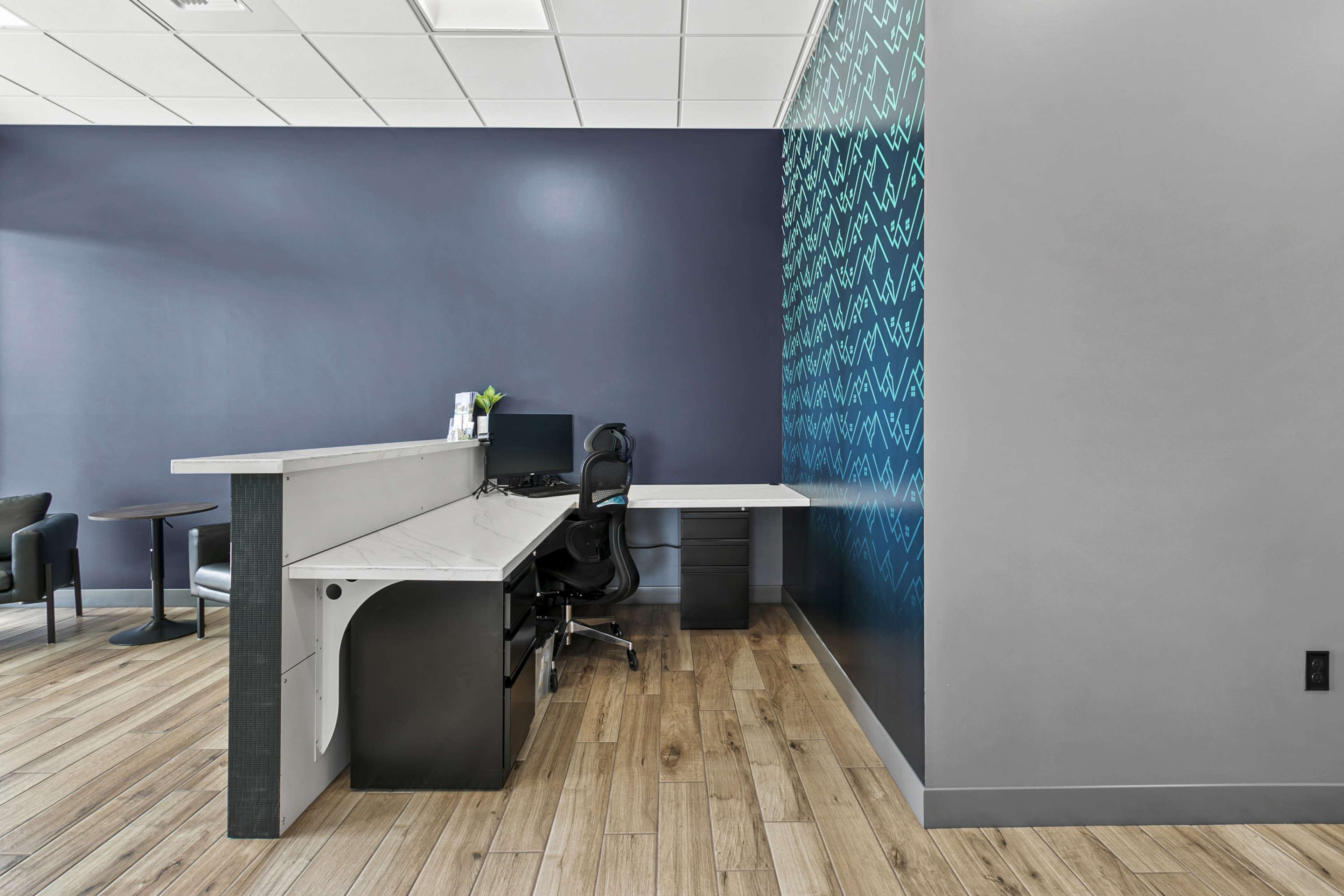 The image shows a modern office reception area with a white counter, a computer setup, a black chair, and a patterned blue wall.