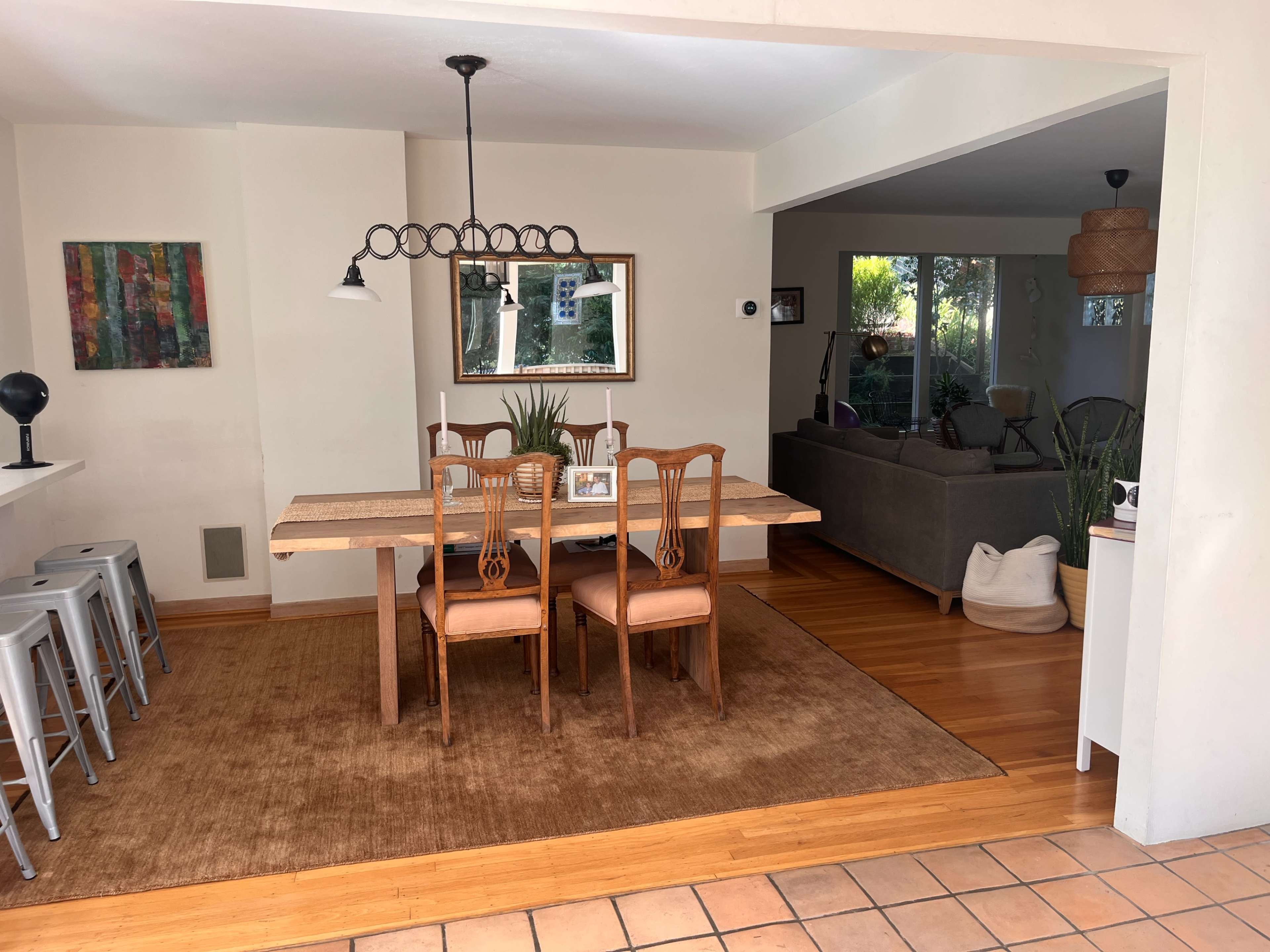 The image shows a dining area with a wooden table and four chairs, accompanied by a rug, while a living room with a gray sofa and plants is visible in the background.