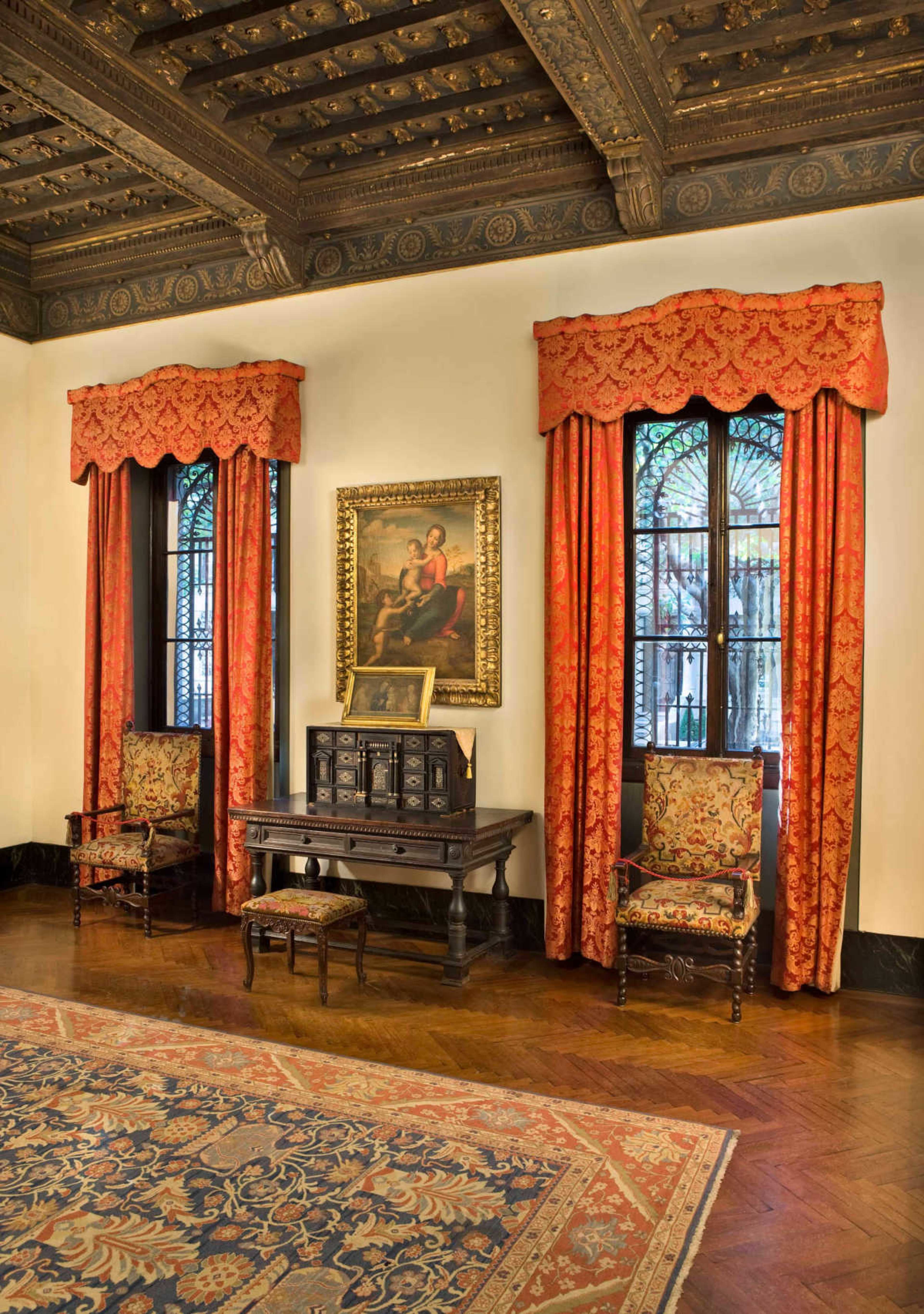 The image depicts an elegantly decorated room featuring two ornate windows with red drapes, two patterned chairs, and an antique table with a decorative cabinet, all set against a richly detailed ceiling.