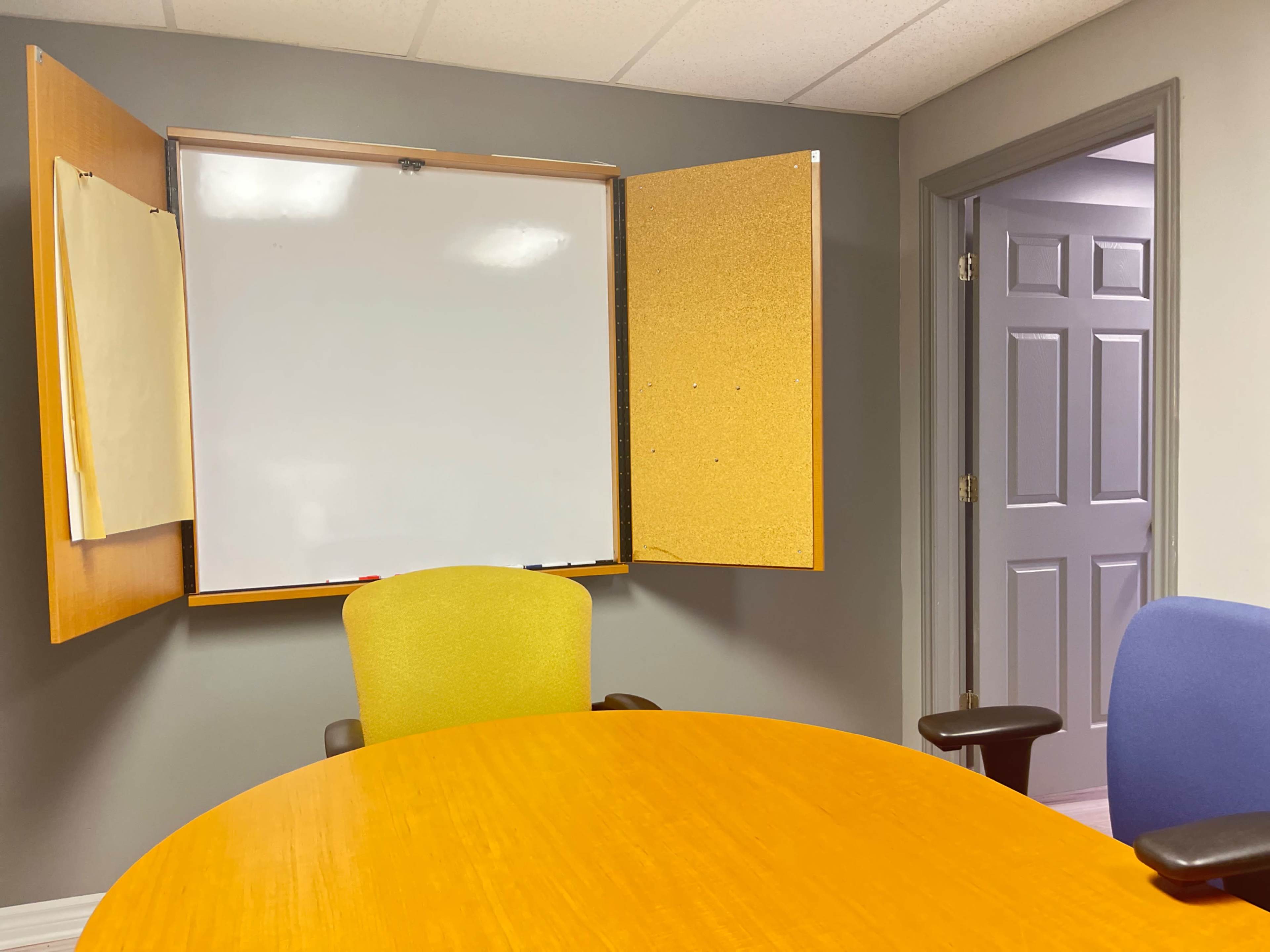 A conference room with a round wooden table, two chairs, a whiteboard mounted on the wall, and a door leading to another room.