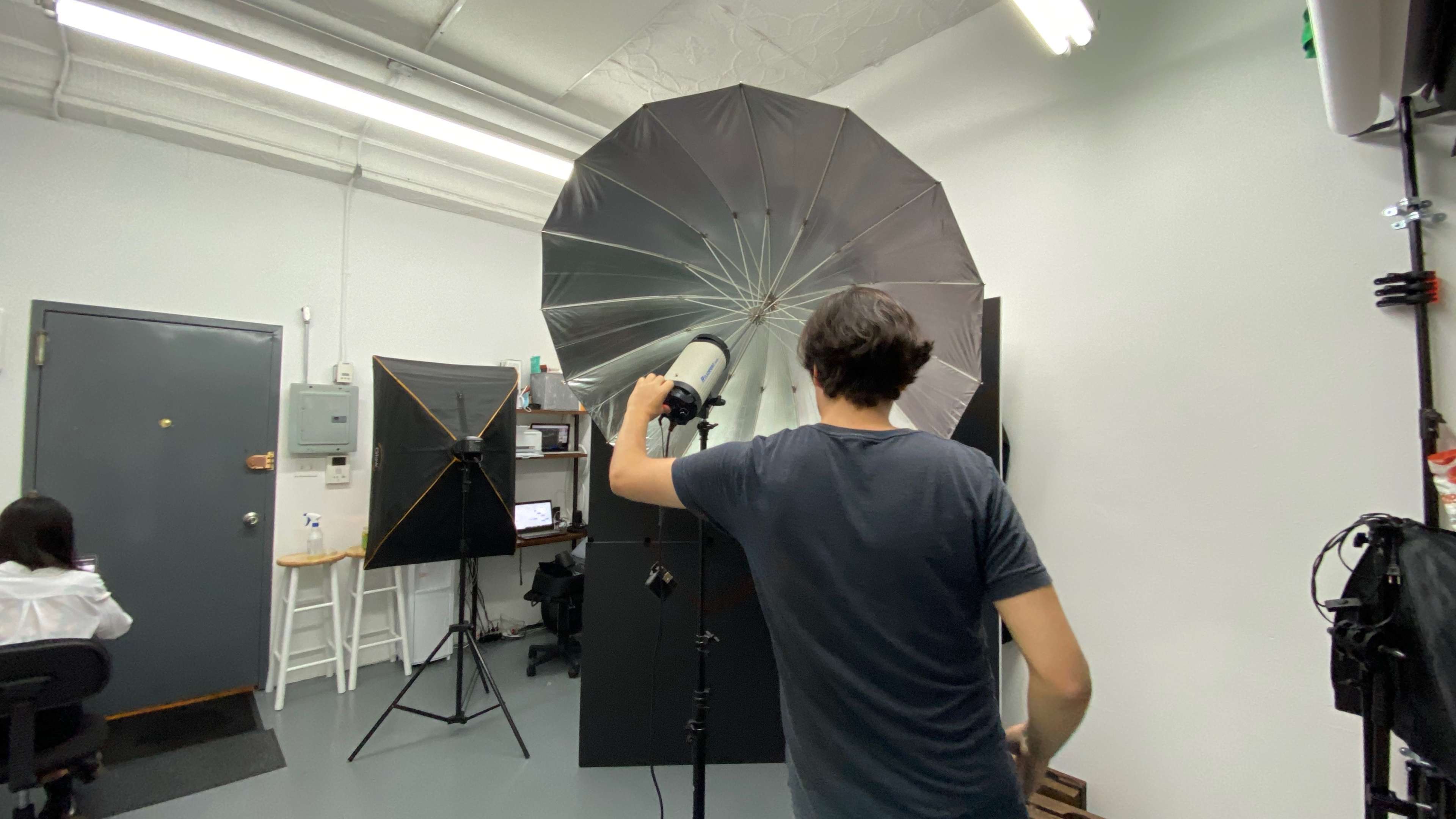 A person stands in a photography studio, adjusting an umbrella light while facing a black backdrop and several pieces of equipment.
