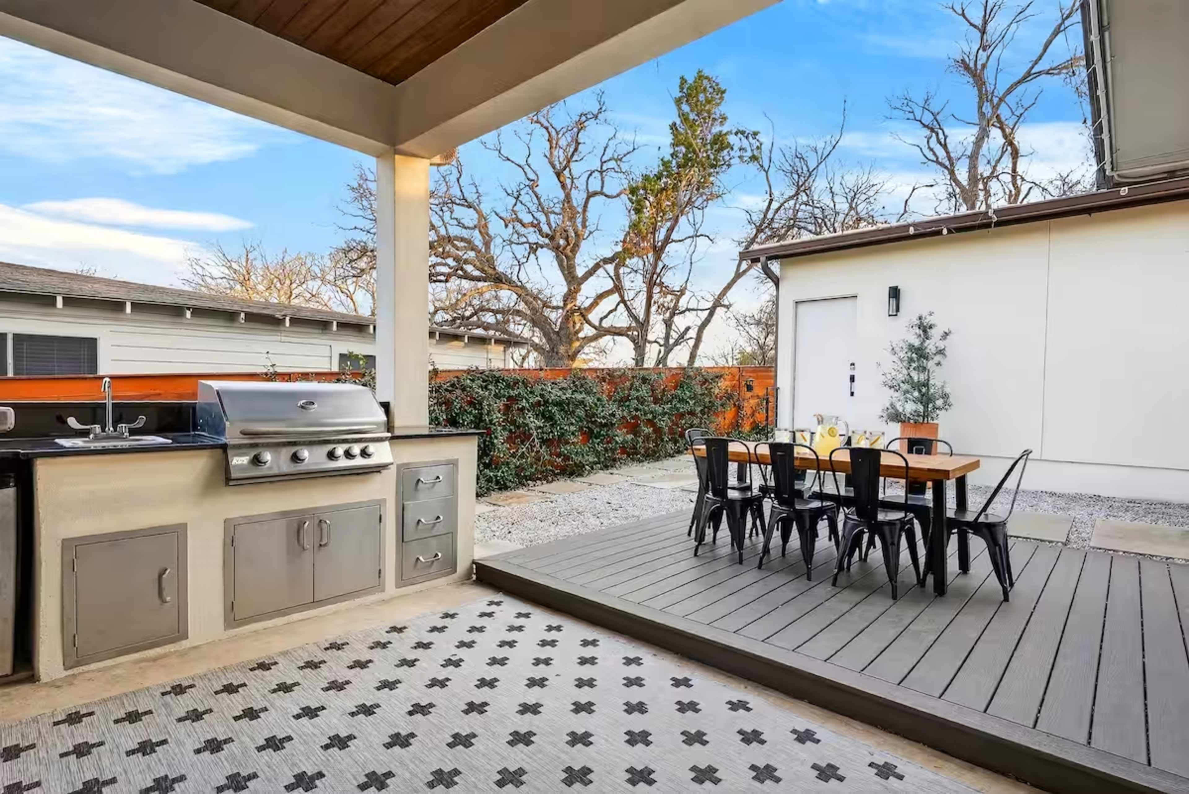 The image shows an outdoor kitchen area featuring a grill and a dining table set against a backdrop of a wooden fence and trees.