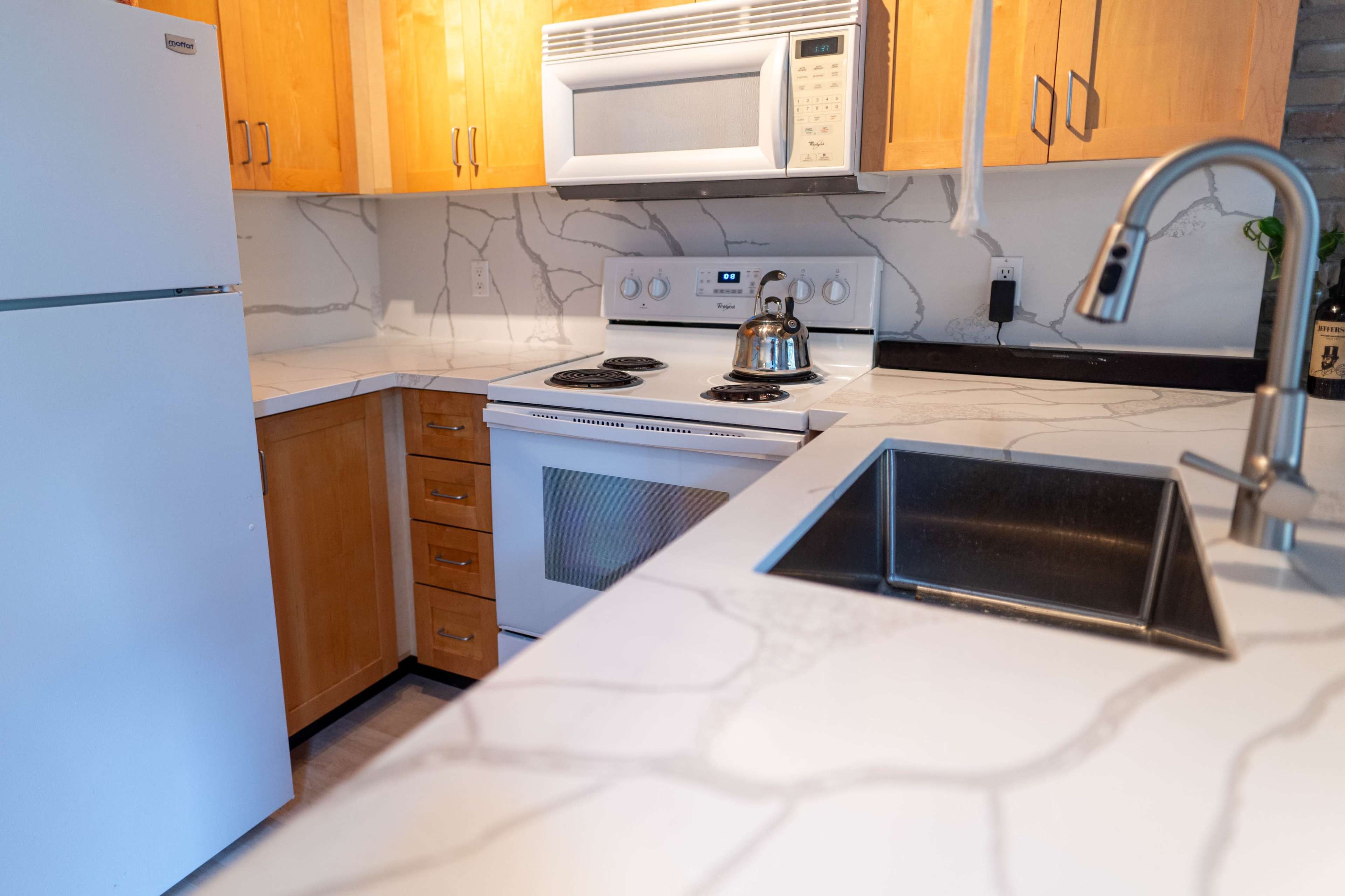 A modern kitchen featuring a white refrigerator, an oven, and a stovetop, all set against a backdrop of light wood cabinetry and a marbled countertop.