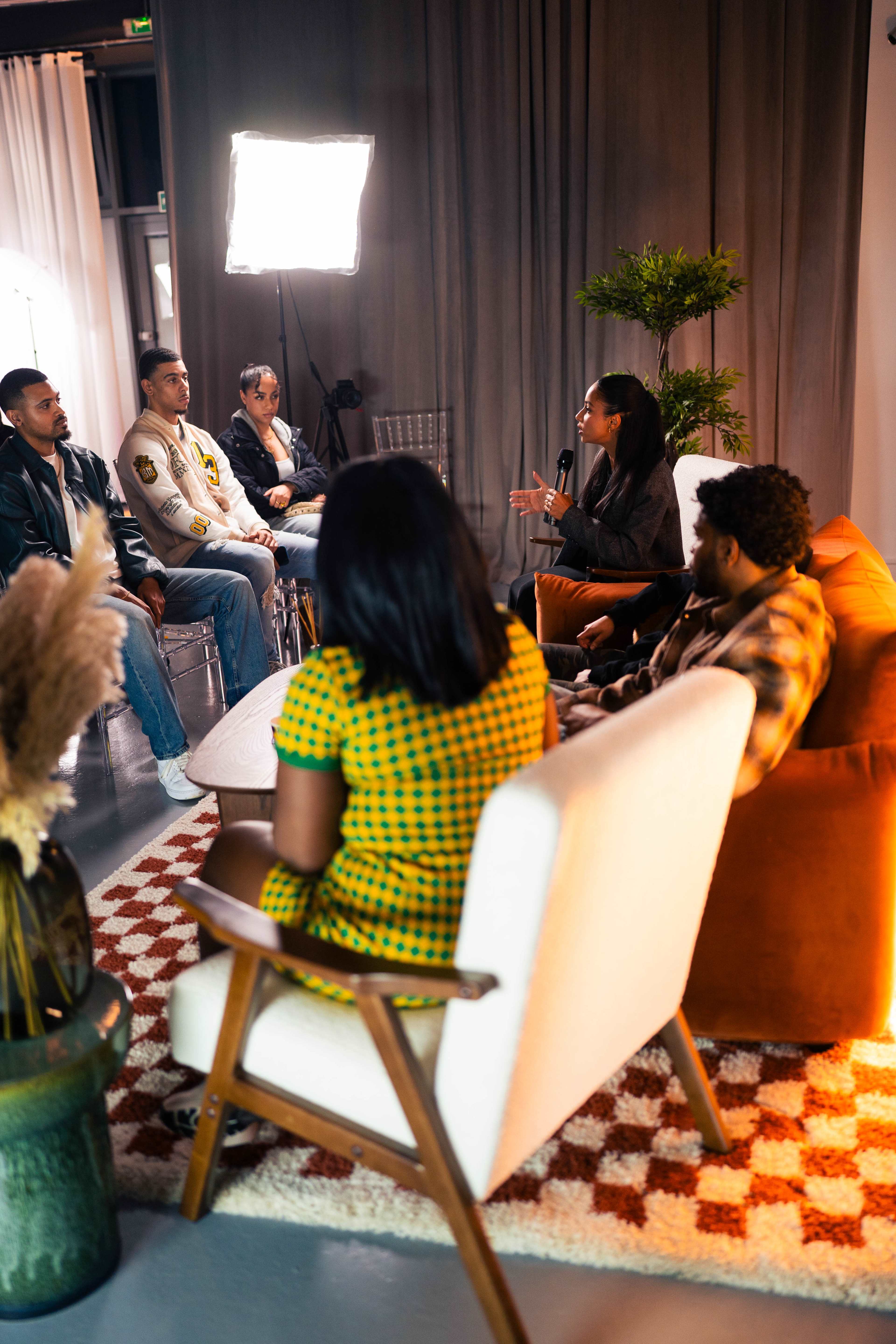 A group of five individuals is seated in a well-lit room, engaged in conversation, while a woman in a checkered dress speaks to them.