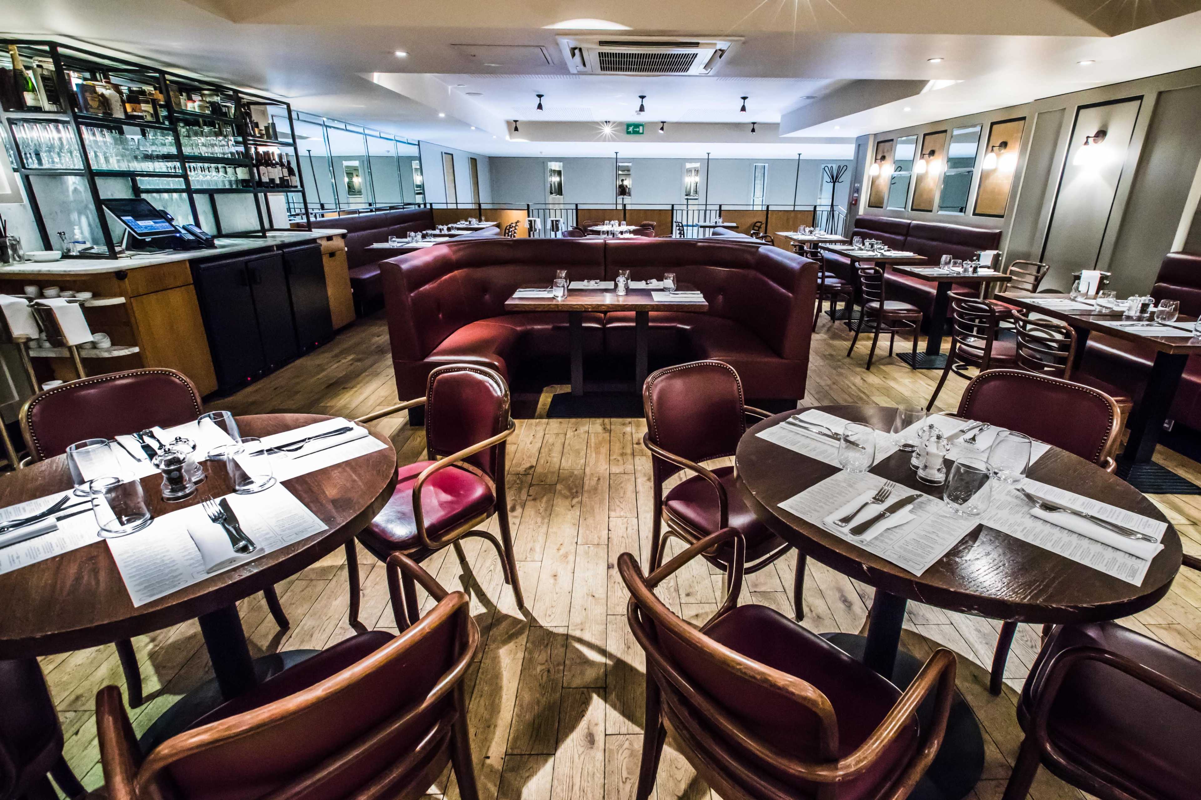 The image shows a partially empty restaurant with wooden floors, round tables set with silverware, and plush booths lining the walls.
