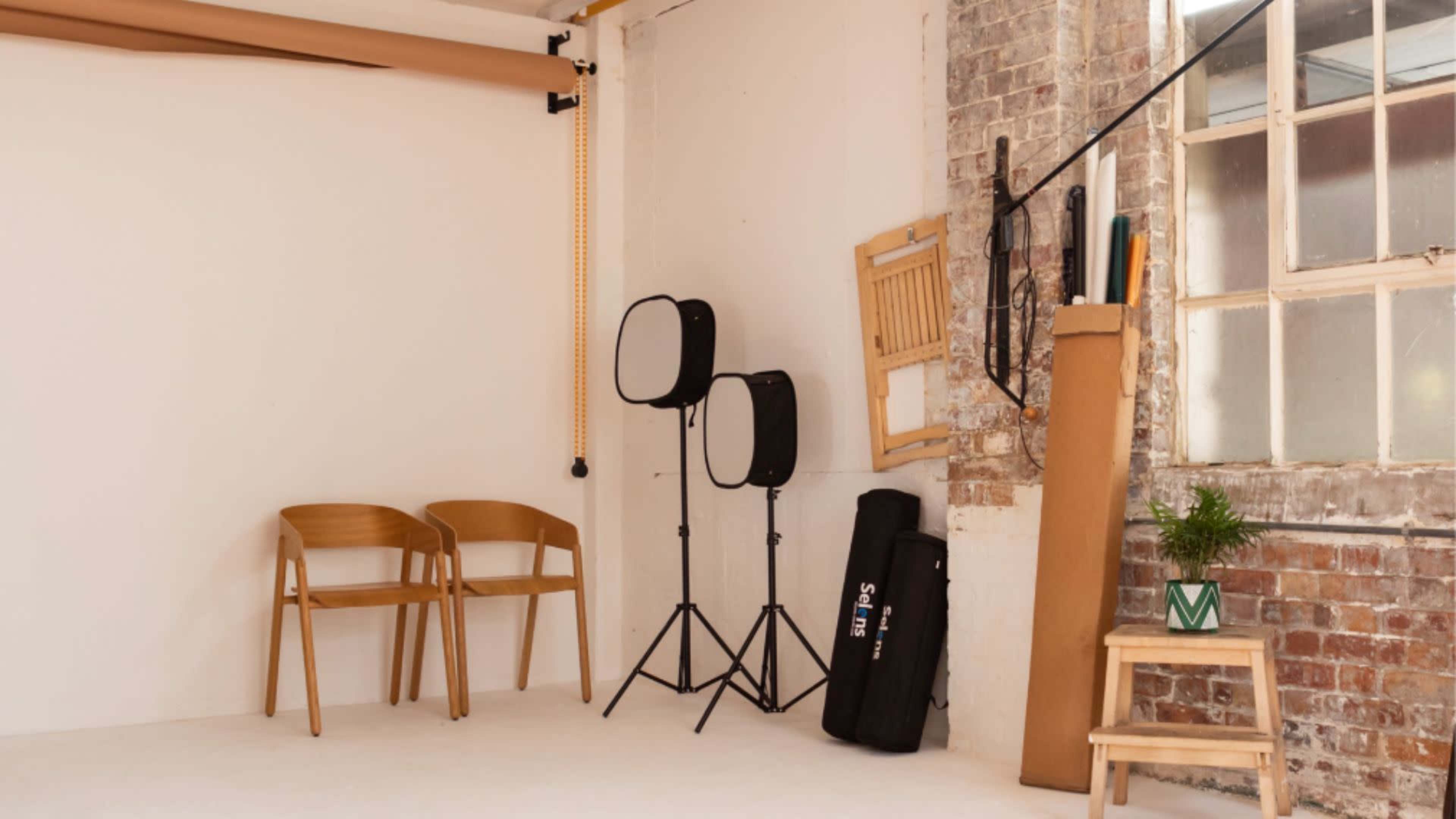 The image shows a photography studio with two wooden chairs, two softbox lights on stands, and various props including a cardboard tube and a small potted plant against a brick wall.