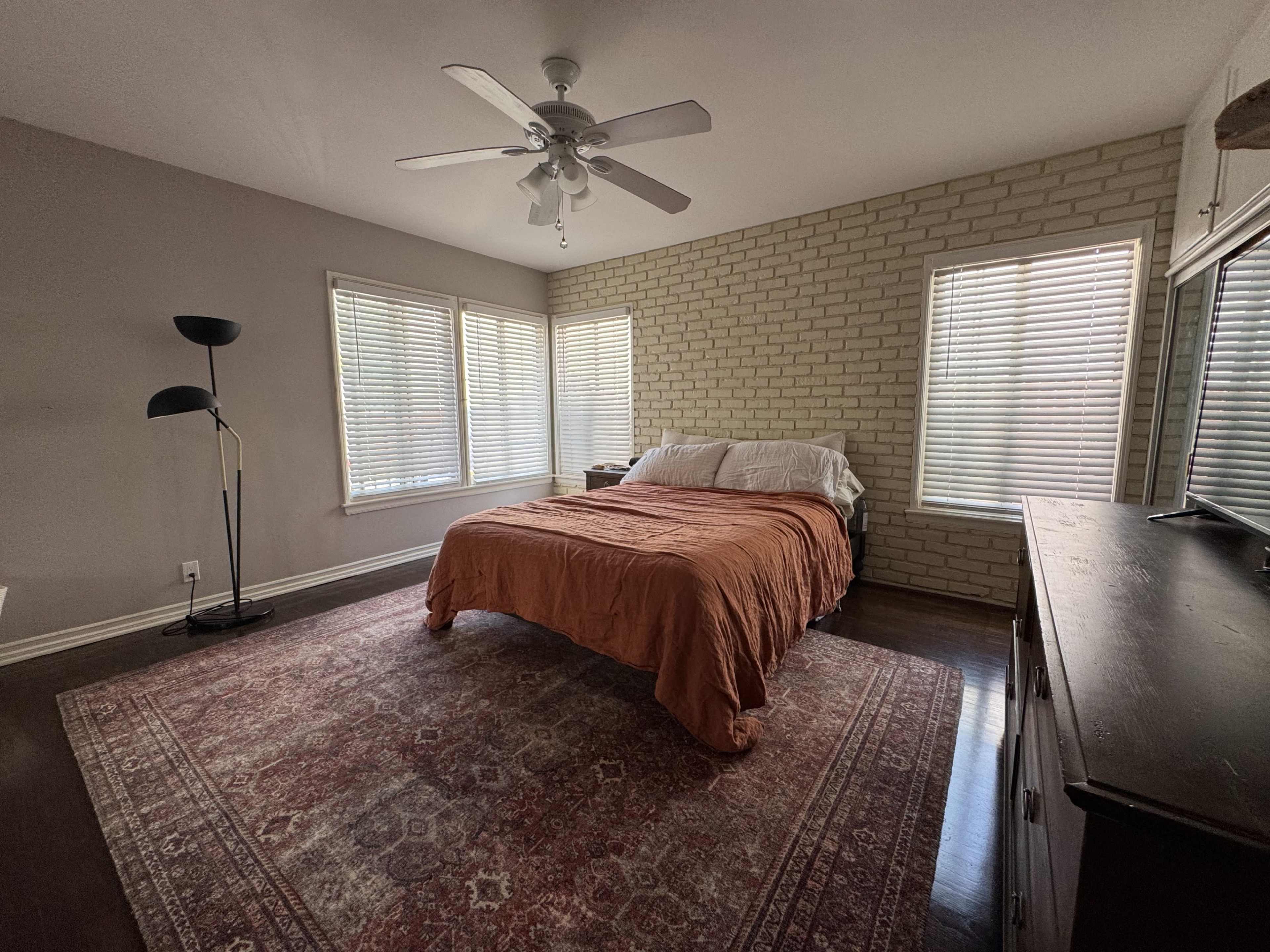 The image shows a bedroom with a bed covered in an orange blanket, a large rug on the floor, and multiple windows with blinds letting in natural light.