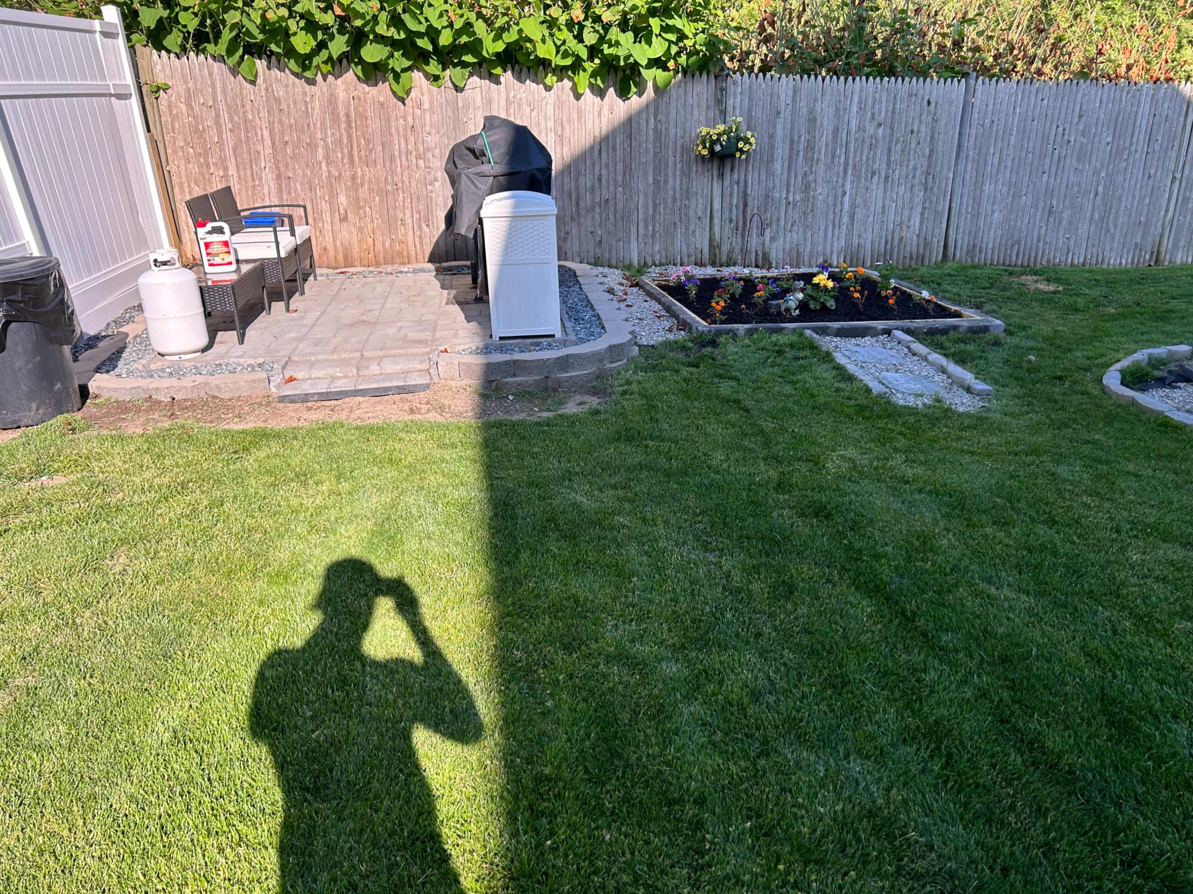 A grassy yard features a patio area with chairs, a grill, and a flowerbed bordered by stones, with a shadow of a person visible in the foreground.