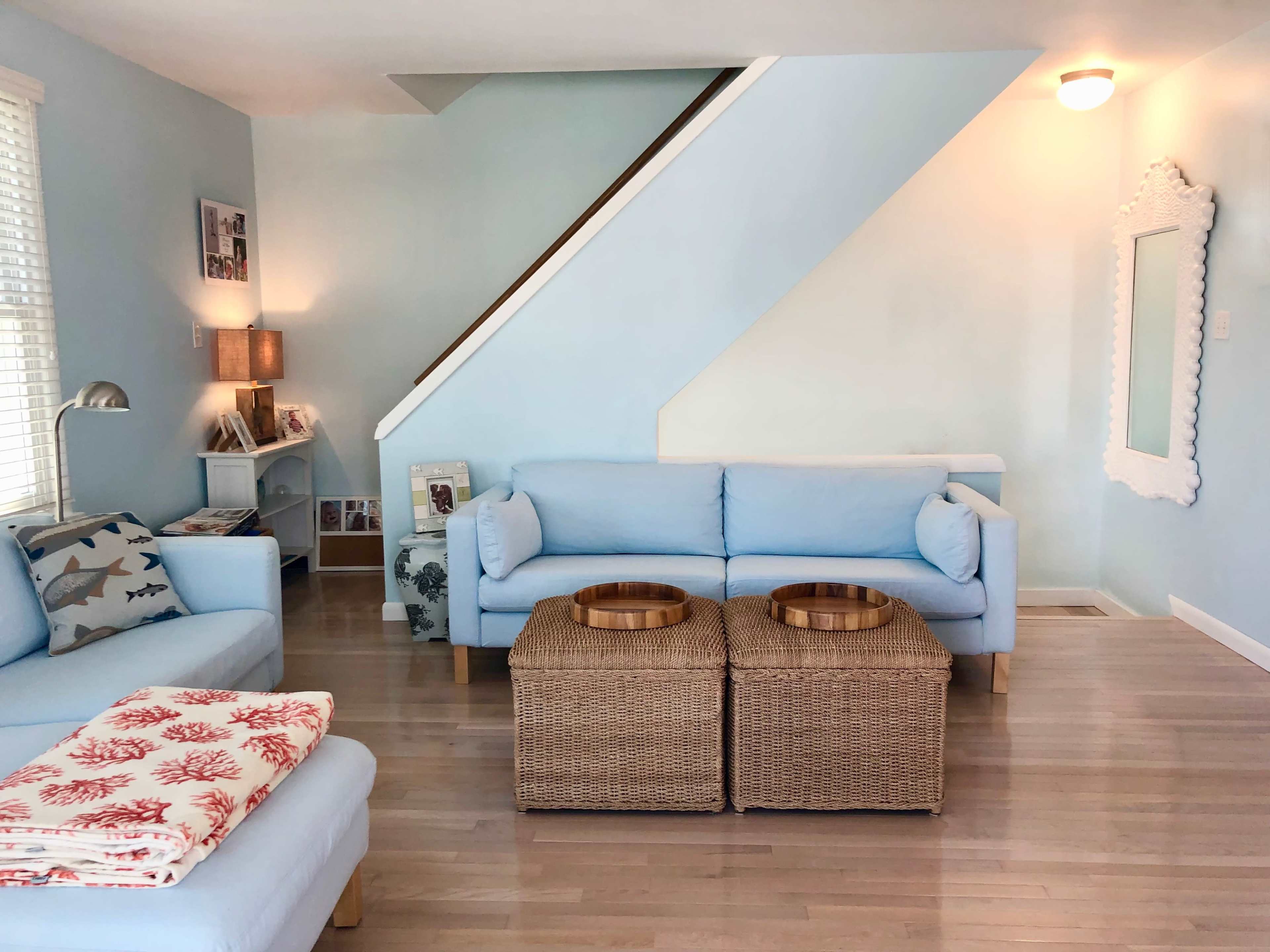 A living room features light blue walls, a staircase, two wicker coffee tables, and soft sofas arranged around a cozy seating area.