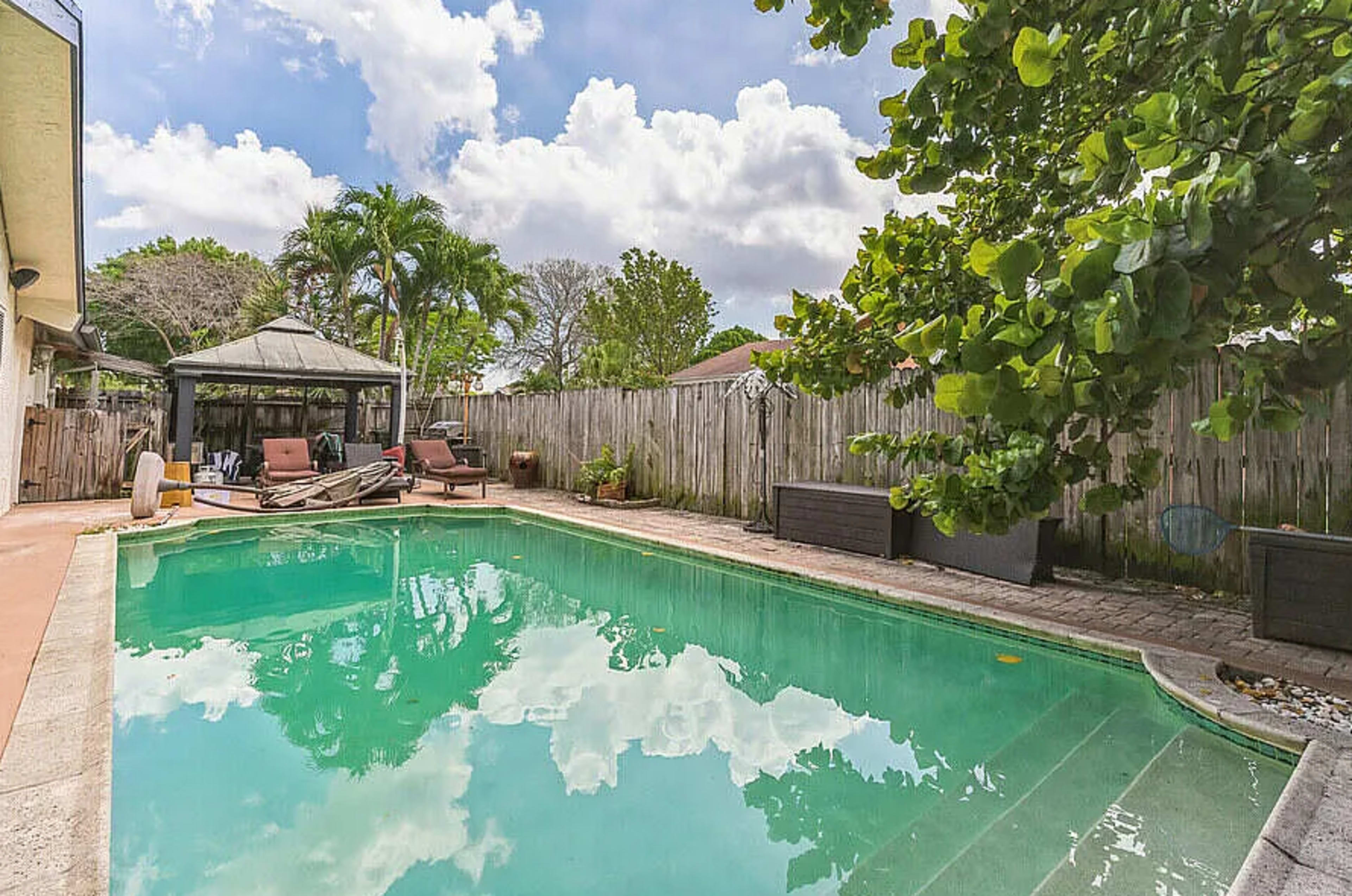 A swimming pool with clear water reflects the sky and surrounding trees, bordered by a wooden fence and patio furniture.
