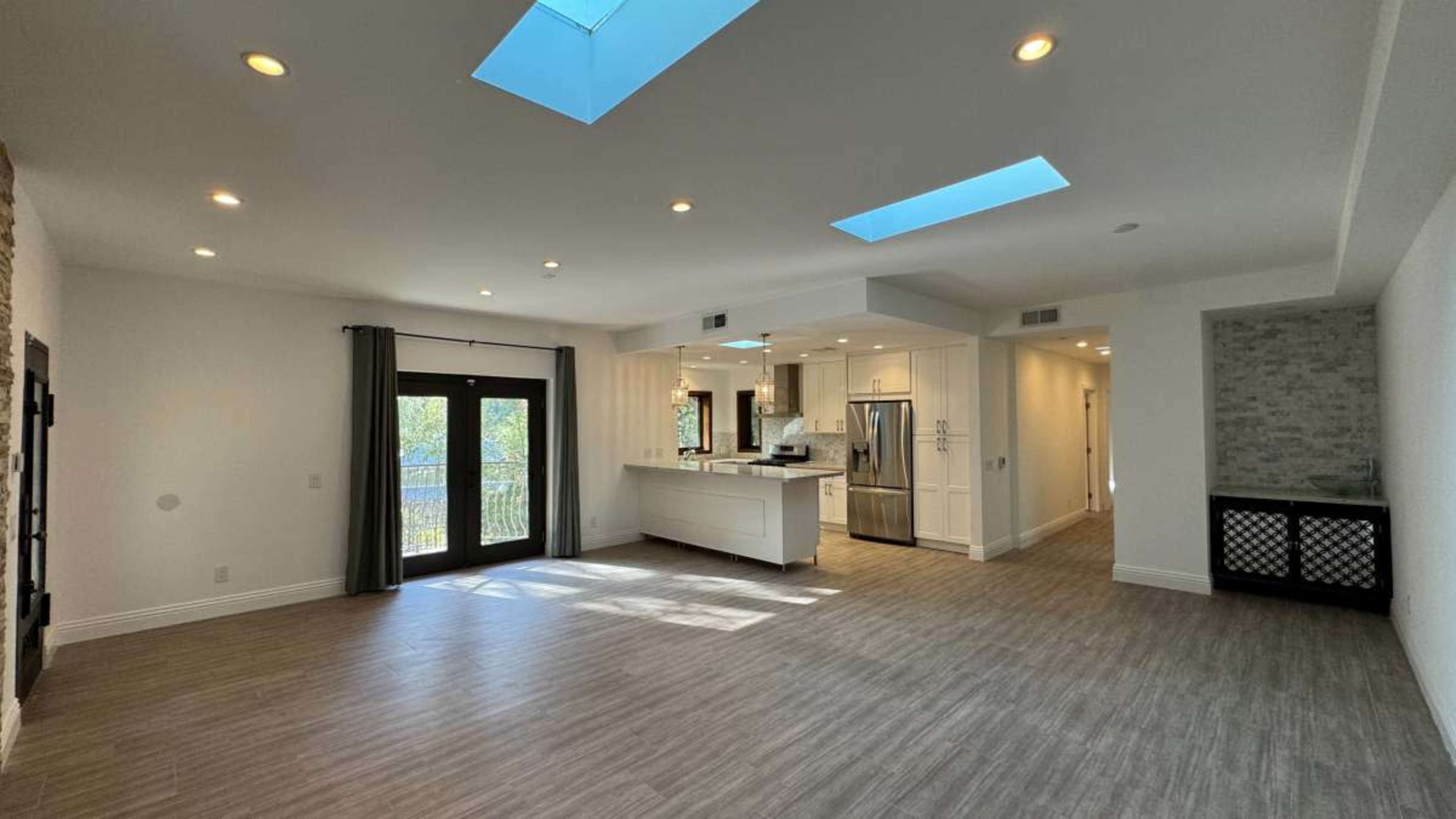 The image shows a spacious, well-lit living area with large skylights, hardwood flooring, and an open kitchen layout.