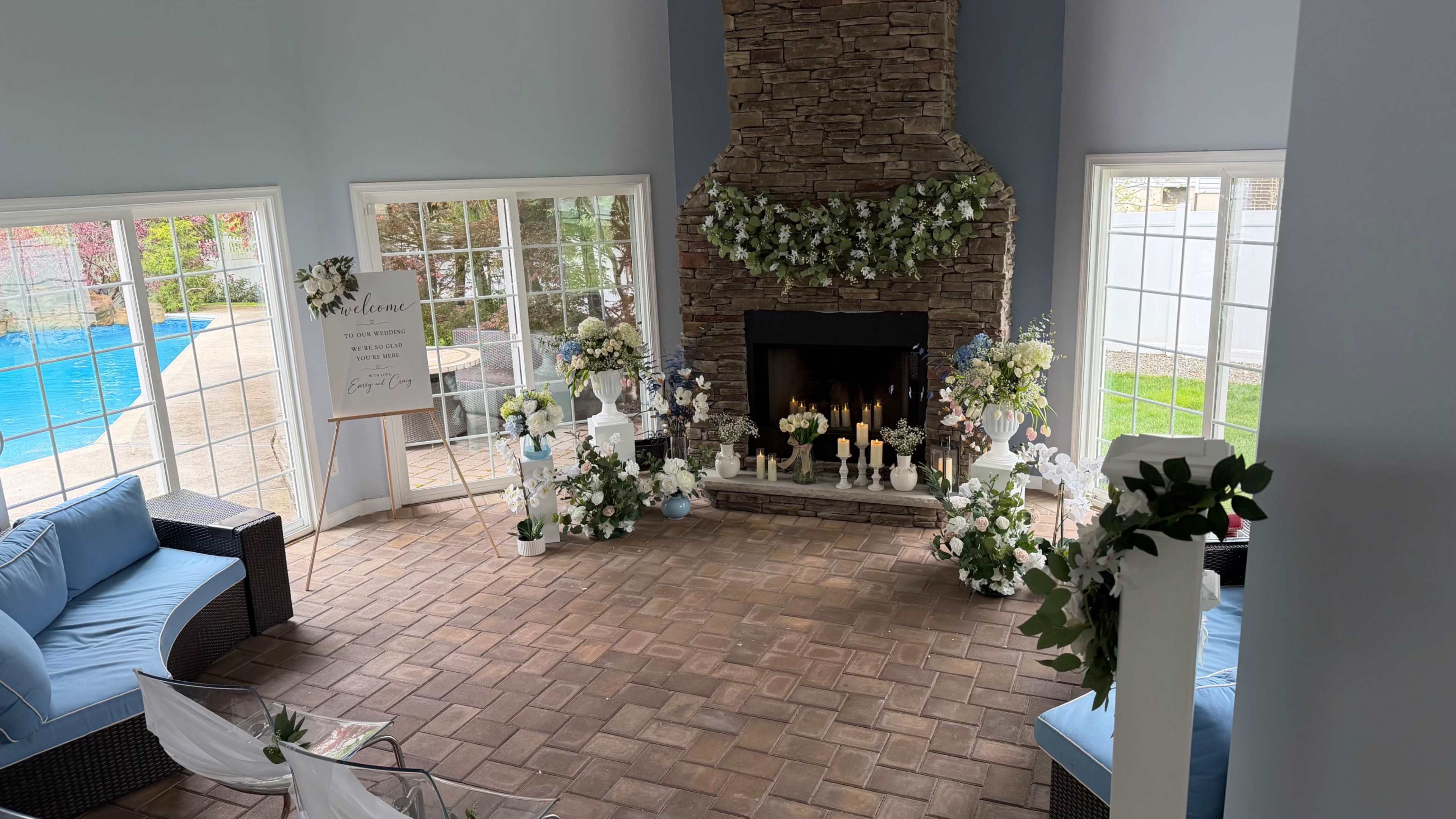 The image shows a well-decorated seating area with a stone fireplace, surrounded by floral arrangements and a swimming pool visible through large windows.