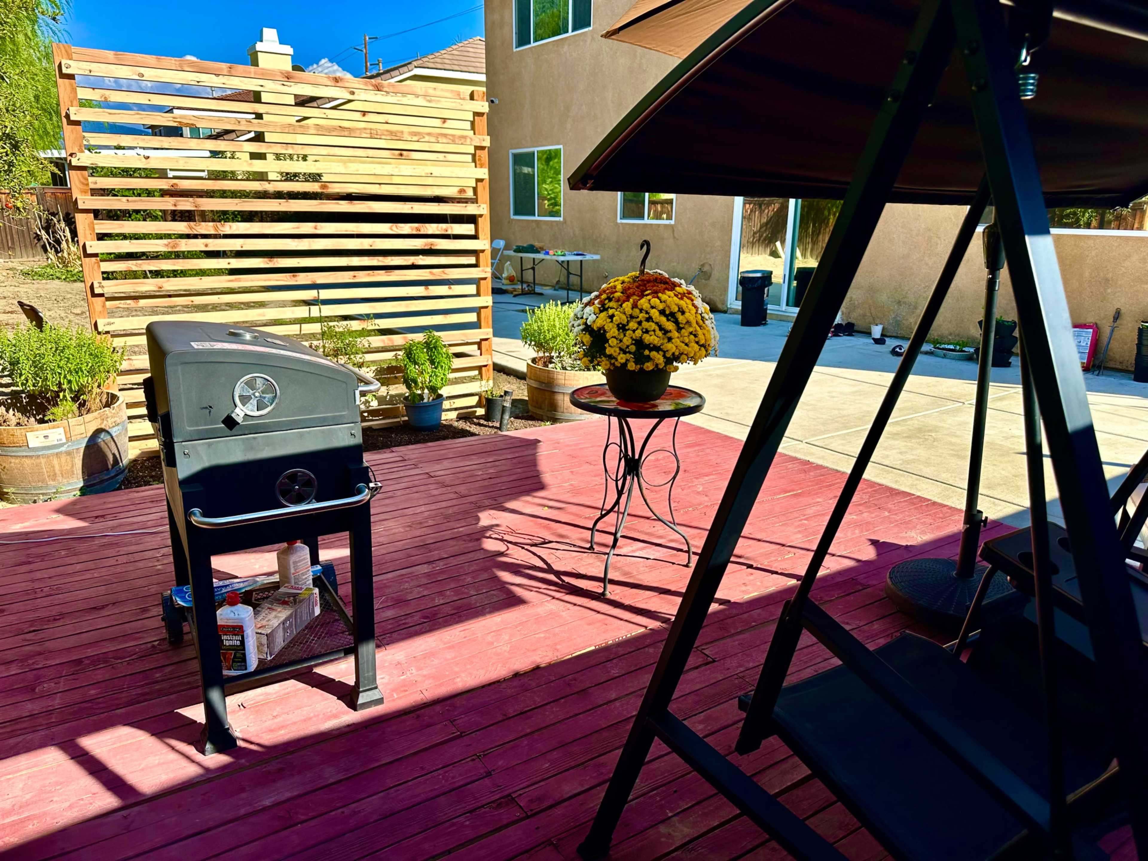 The image shows a patio area with a black grill, a table with a pot of yellow flowers, and a seating area under a swing canopy, surrounded by wooden fencing and plants.