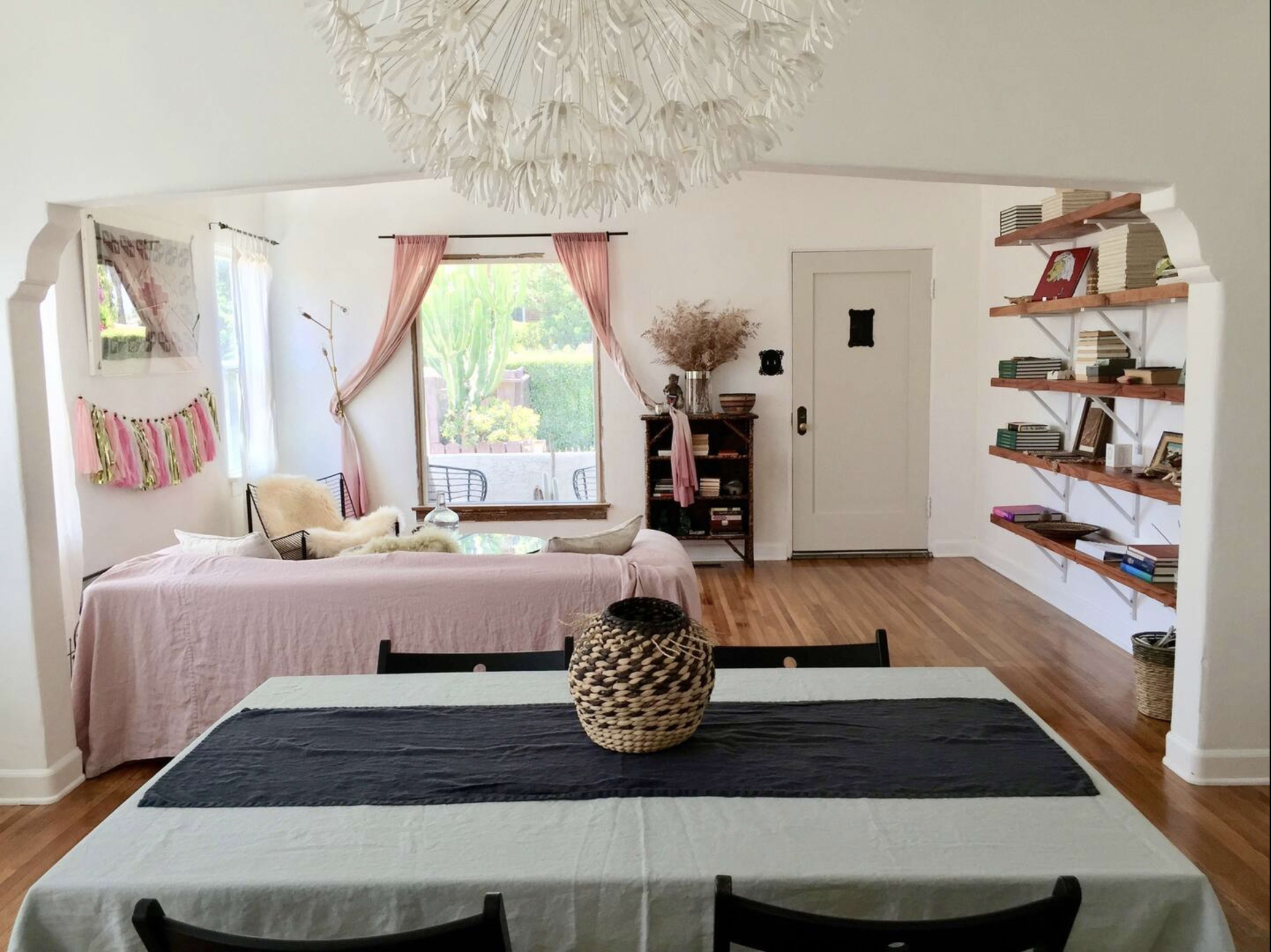 The image depicts a spacious, light-filled room featuring a dining table with a black runner, a couch draped in a pink covering, and a large window with sheer curtains.