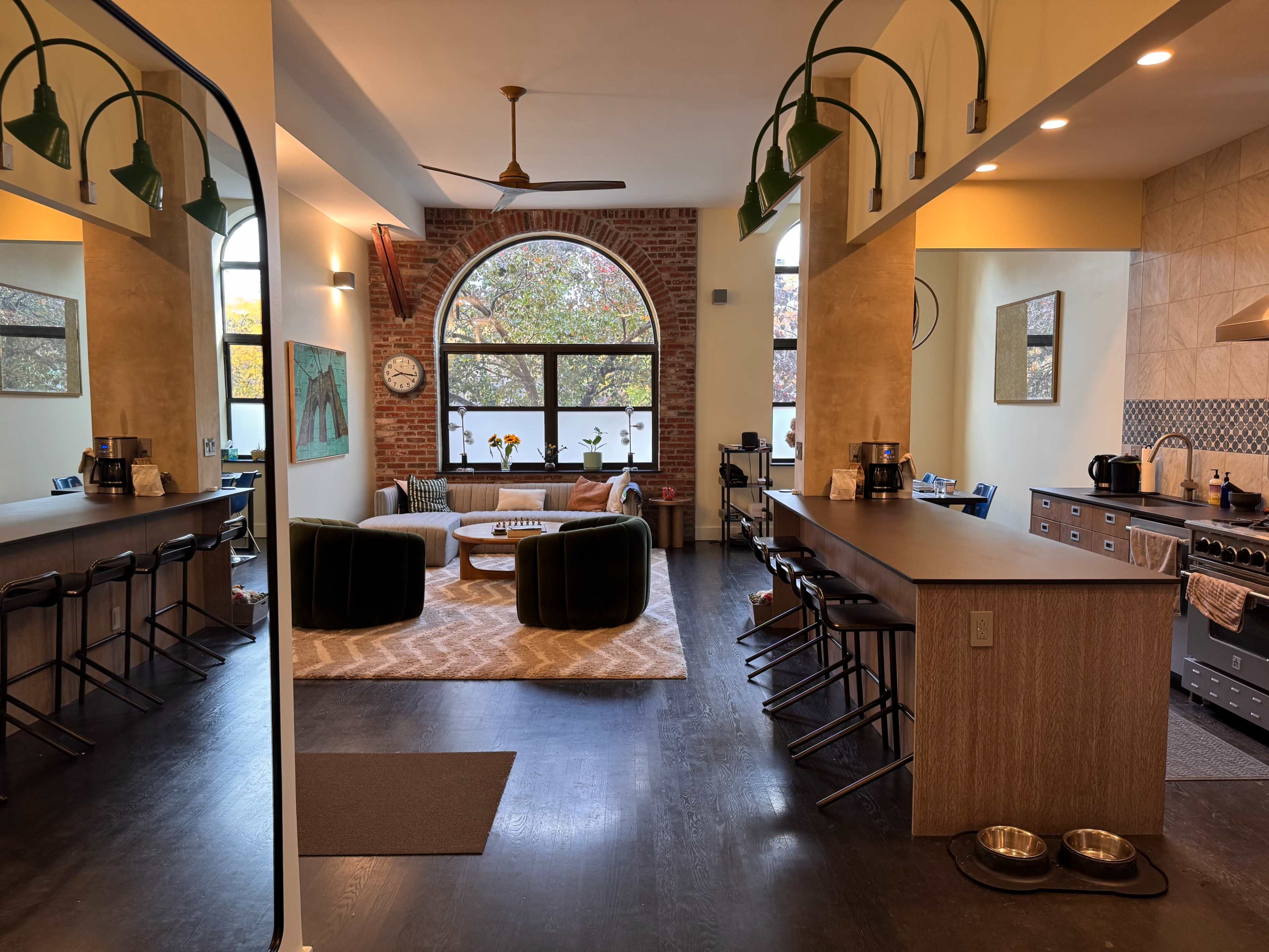 The image shows a spacious, modern kitchen and living area with large windows, a brick accent wall, and a dining area featuring a round table and circular seating.