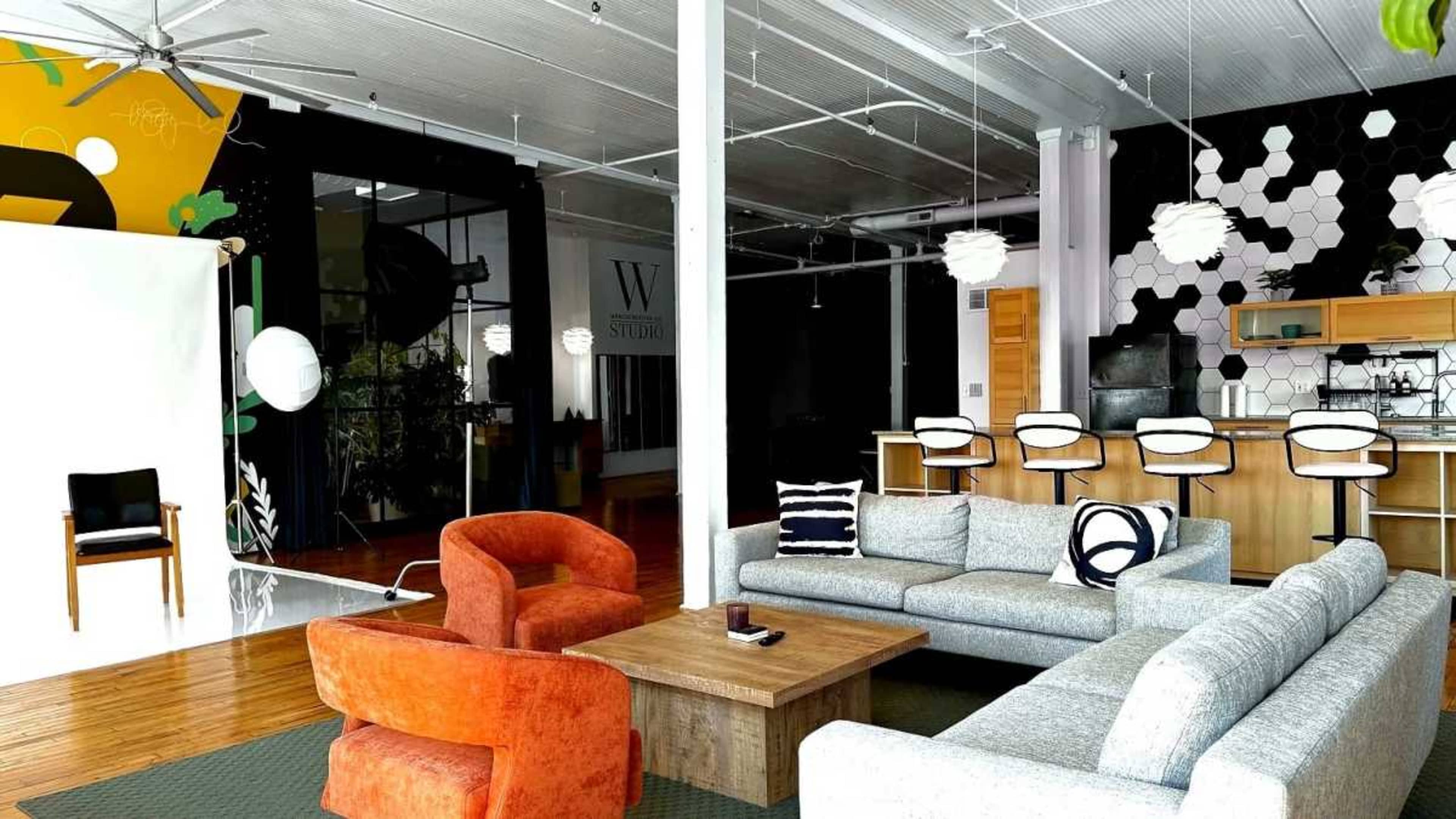 The image shows a spacious modern living area with a gray sofa set, an orange armchair, a wooden coffee table, and a kitchen bar in the background.